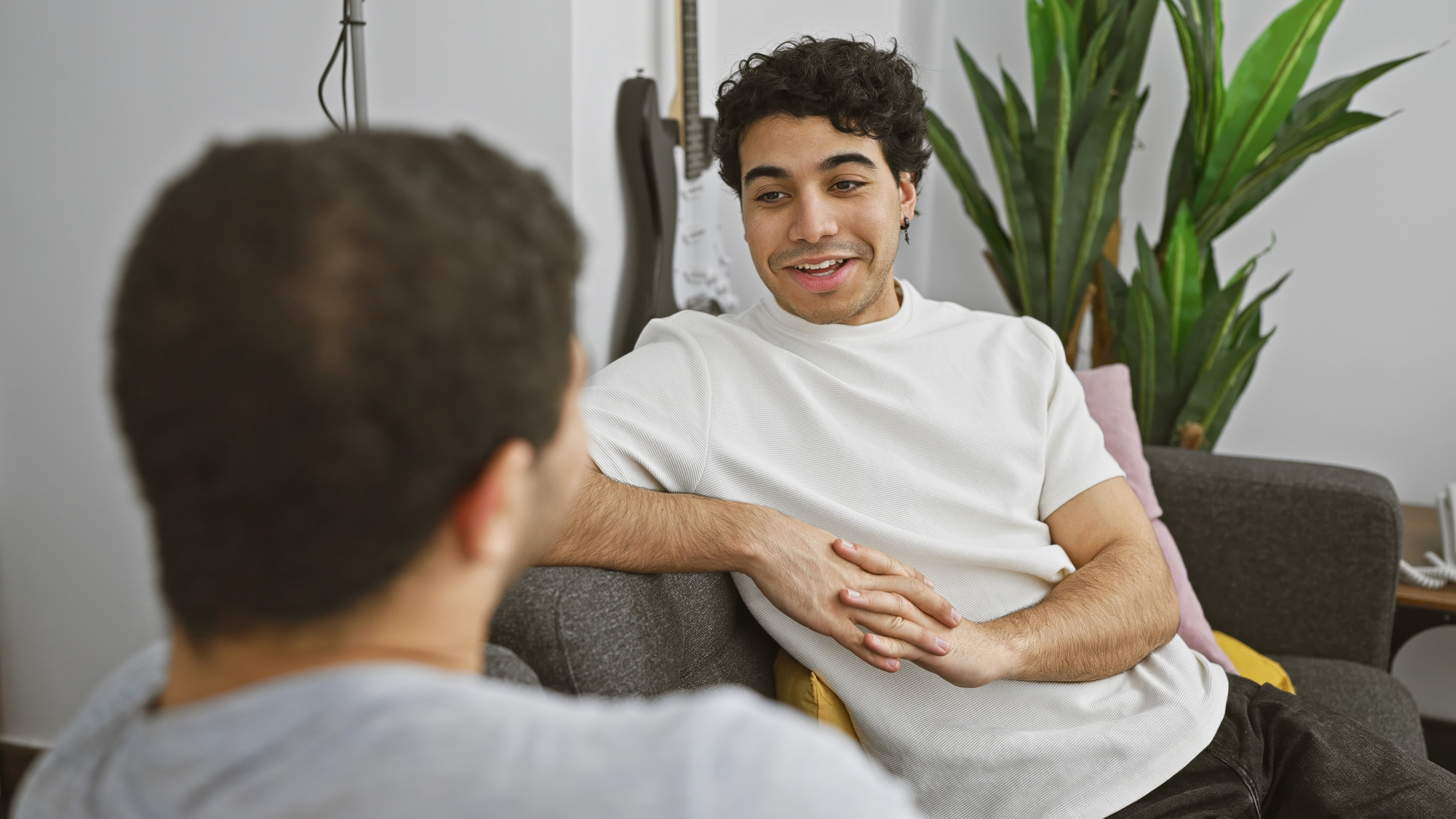 Two people sitting on a couch engage in a conversation in a living room. One person with curly hair wears a white shirt, while the other has their back to the camera. A guitar and a tall plant are in the background.