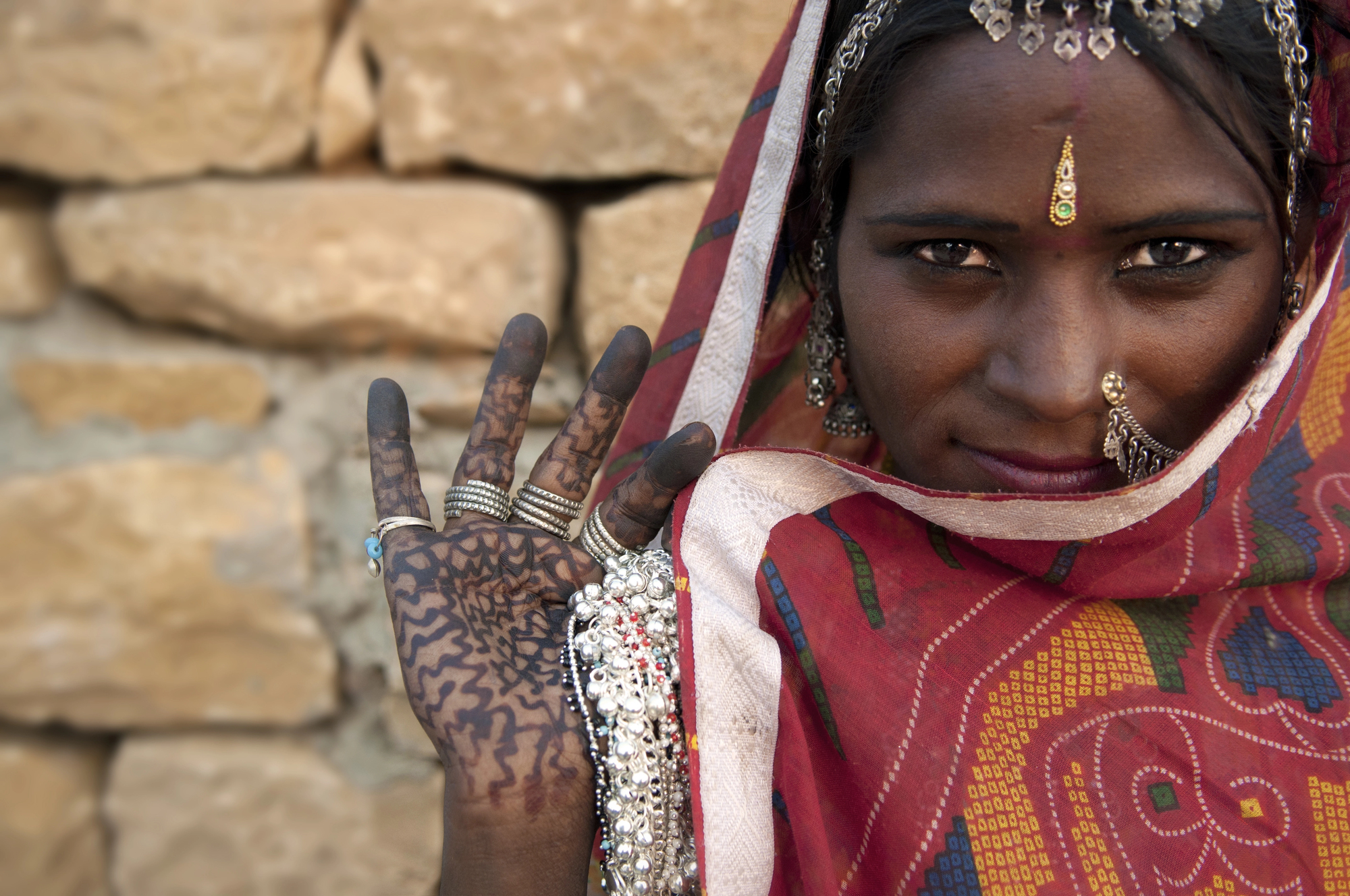 A woman wearing a colorful headscarf adorned with jewelry holds up her hand, displaying intricate henna designs. She stands in front of a beige stone wall, partially smiling and showcasing a nose ring and forehead ornament.