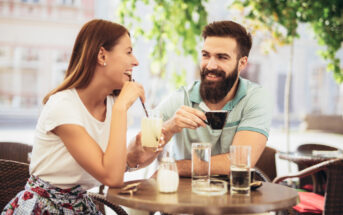 A woman and a man sitting at an outdoor café table, smiling and enjoying drinks. The woman holds a glass with a straw, and the man holds a small cup. They are surrounded by greenery and buildings are visible in the background.