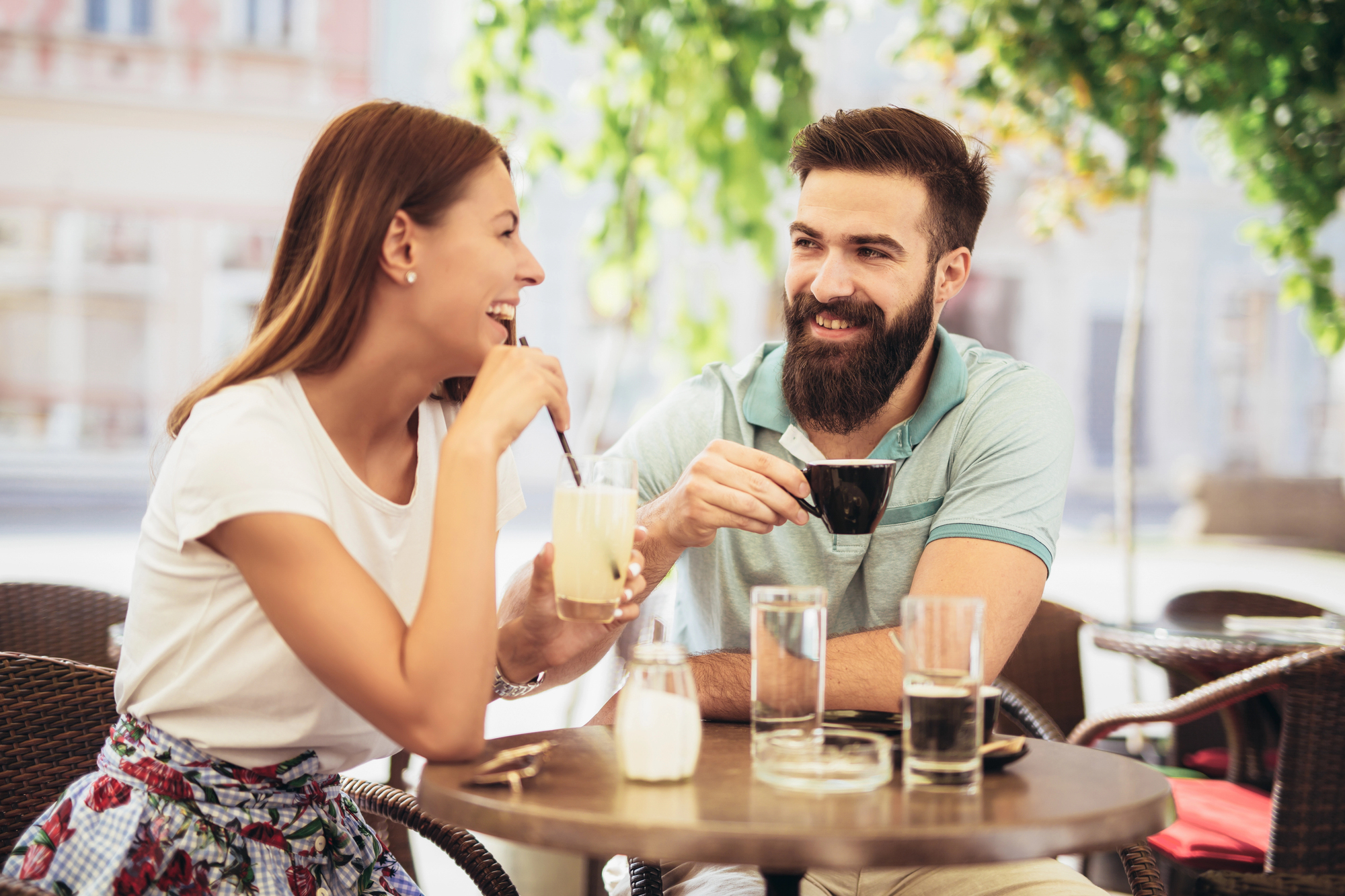 A woman and a man sitting at an outdoor café table, smiling and enjoying drinks. The woman holds a glass with a straw, and the man holds a small cup. They are surrounded by greenery and buildings are visible in the background.