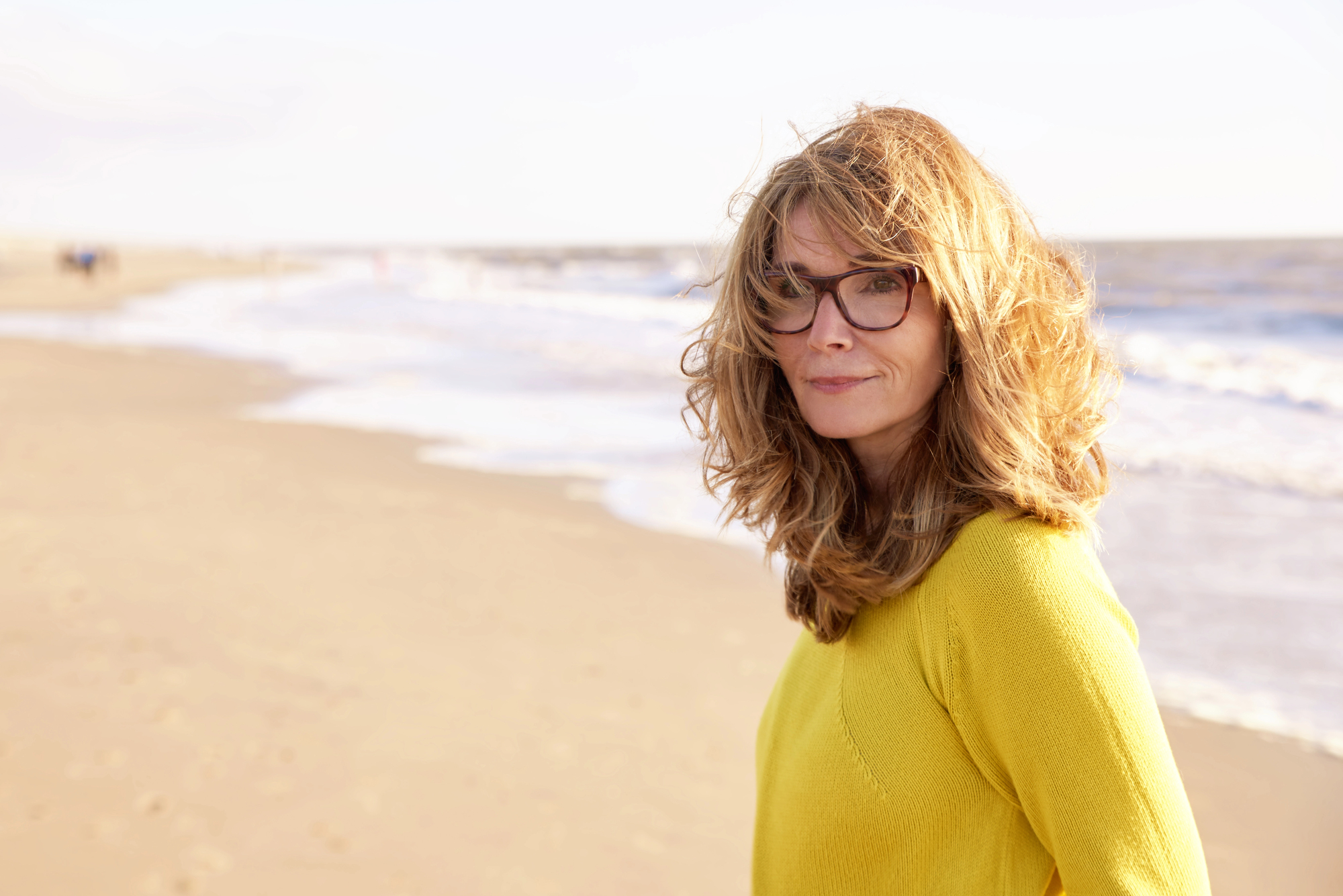 A woman with wavy blonde hair and glasses stands on a sandy beach, wearing a yellow sweater. The ocean waves roll in the background, and the sky is bright and clear.