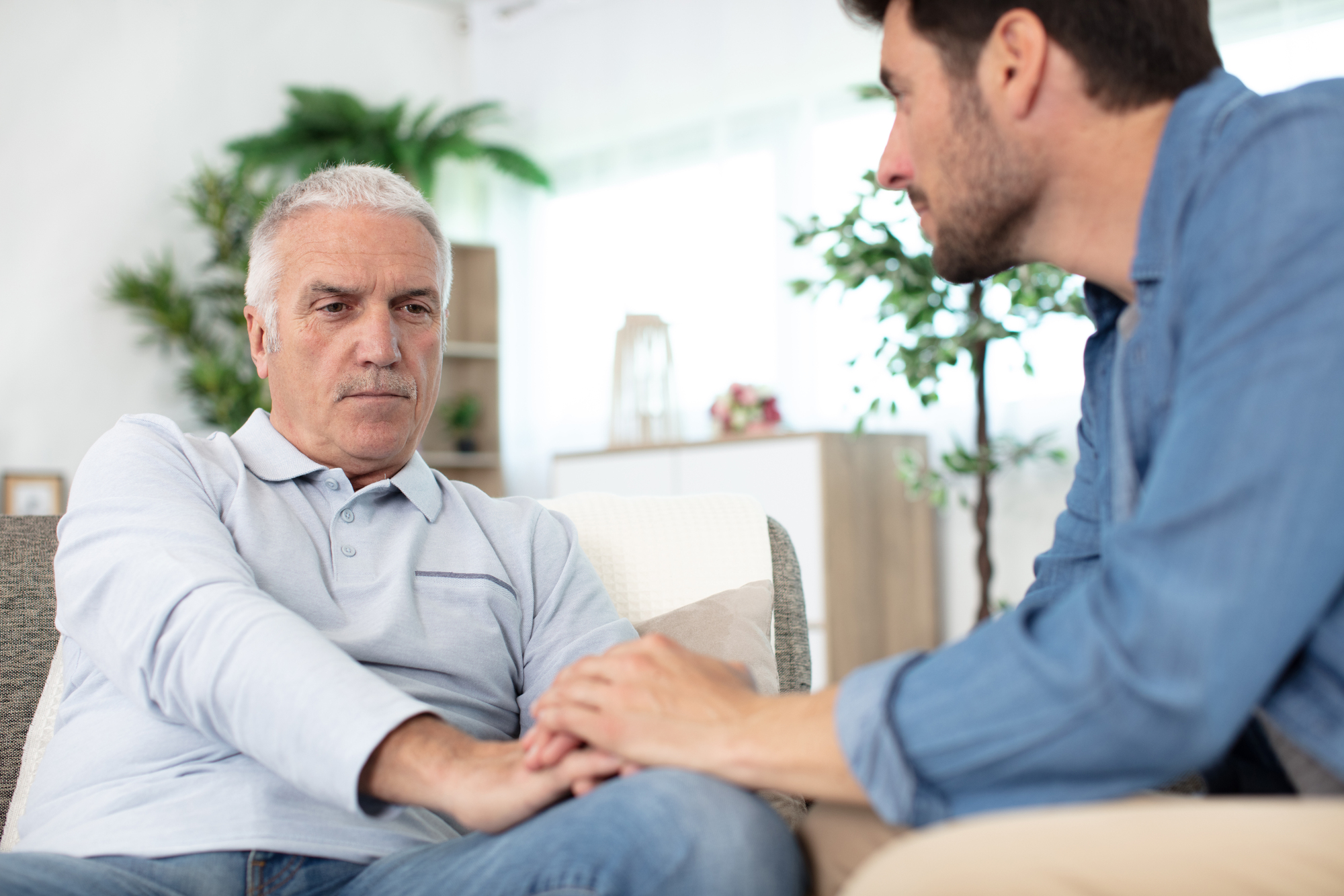 A concerned younger man holds the hands of an older man sitting on a couch in a bright living room. The older man appears serious, while the younger offers comfort. There are houseplants and soft natural light in the background.