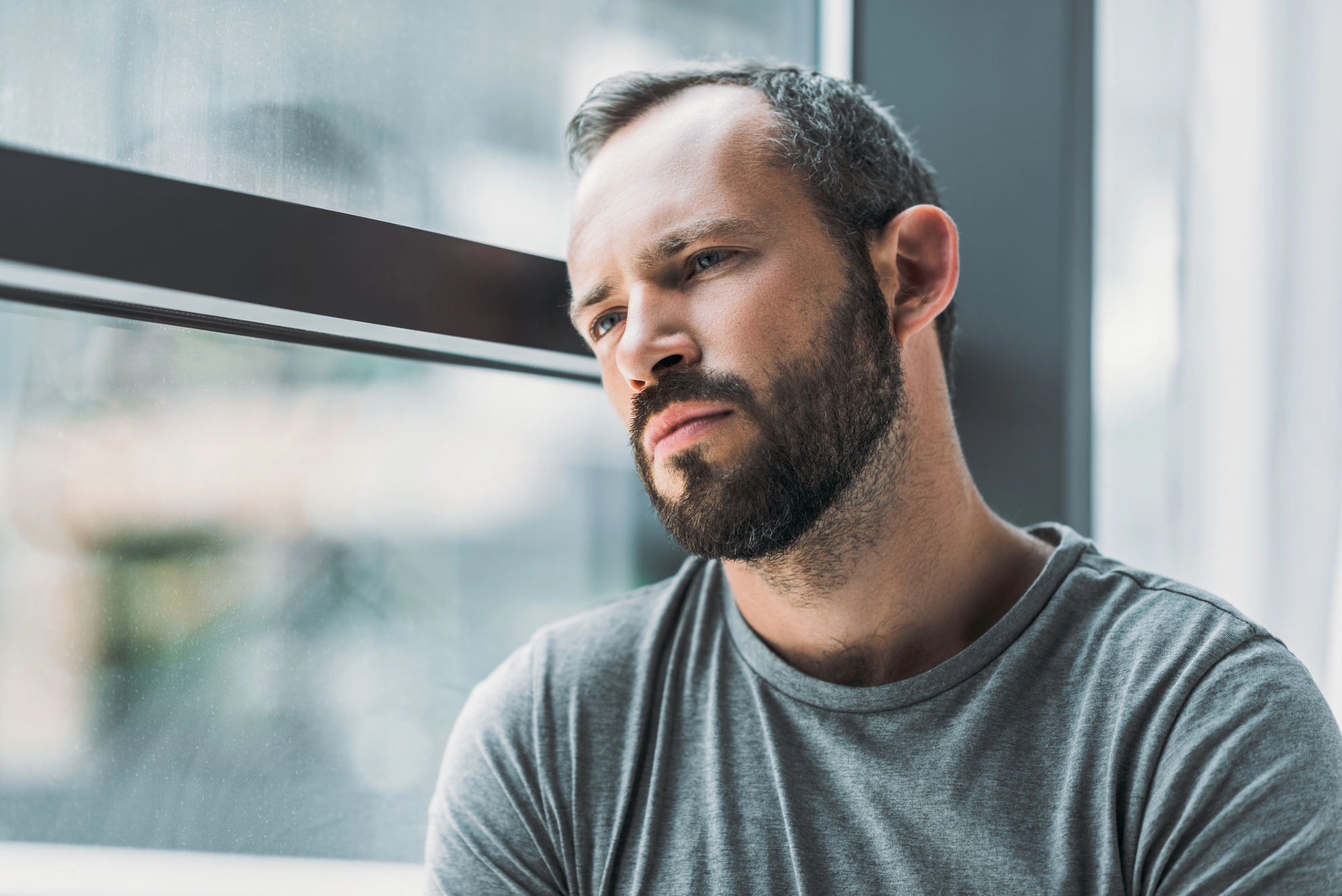 A man with a beard and short hair is leaning against a window, gazing thoughtfully into the distance. He is wearing a gray T-shirt and appears to be lost in deep contemplation. The lighting is soft and natural.