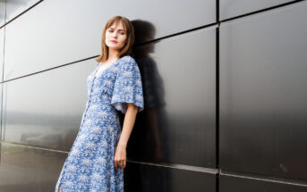 A woman in a blue floral dress leans against a dark, reflective wall. She has shoulder-length hair and looks towards the camera with a calm expression. The setting appears modern and urban.