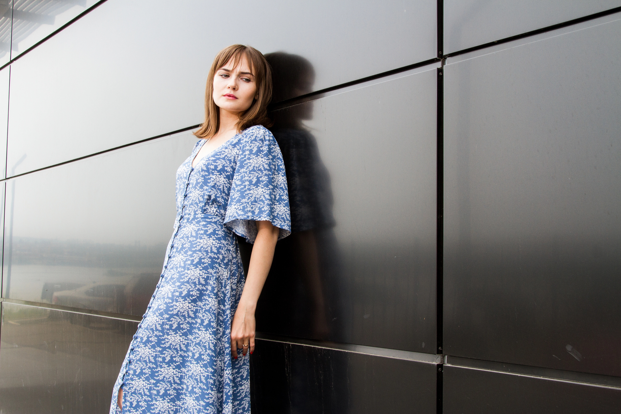 A woman in a blue floral dress leans against a dark, reflective wall. She has shoulder-length hair and looks towards the camera with a calm expression. The setting appears modern and urban.