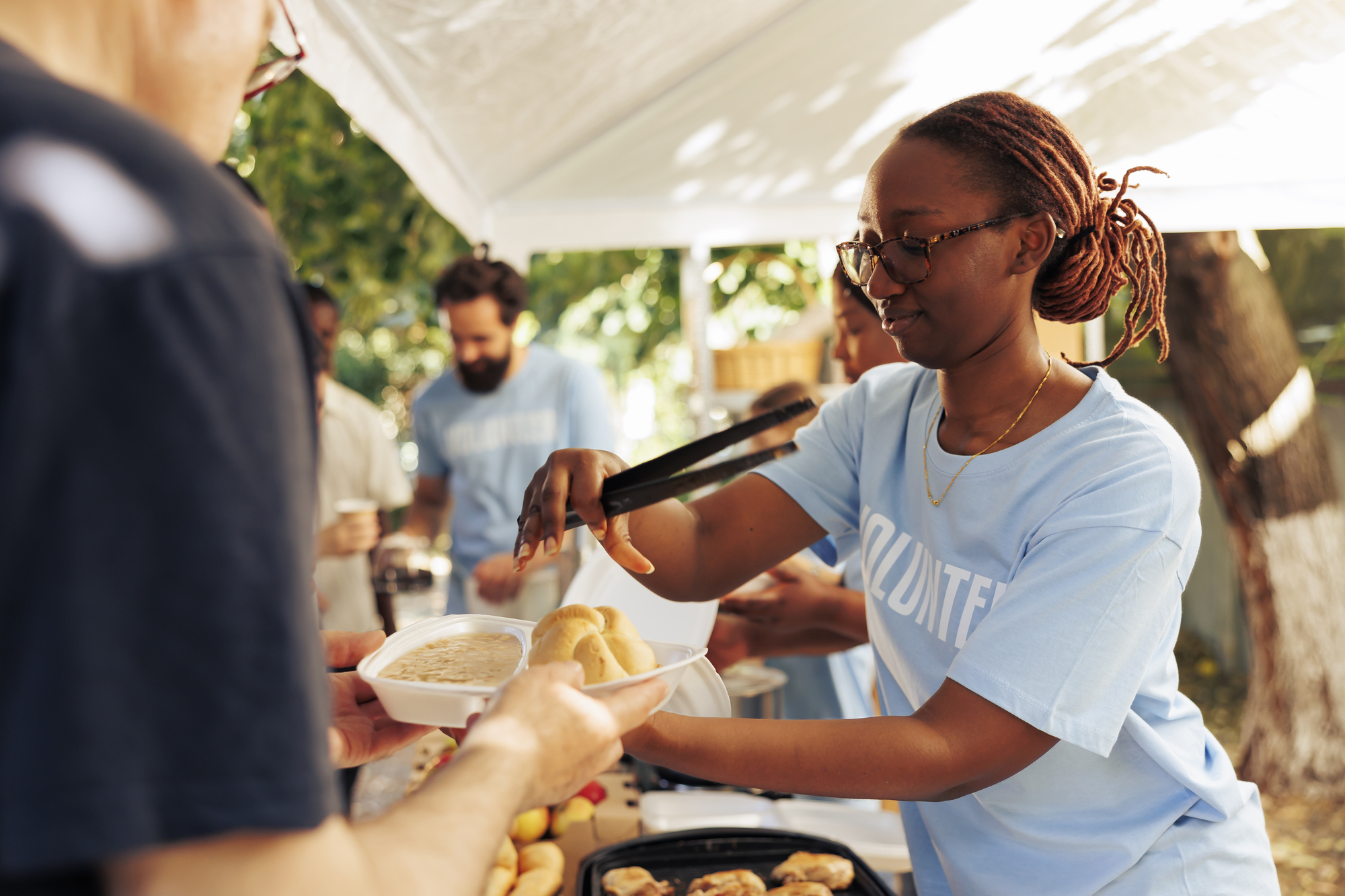 A volunteer with glasses serves food to a person at an outdoor community event. She uses tongs to place food in a container under a canopy. Other volunteers and attendees are visible in the background.