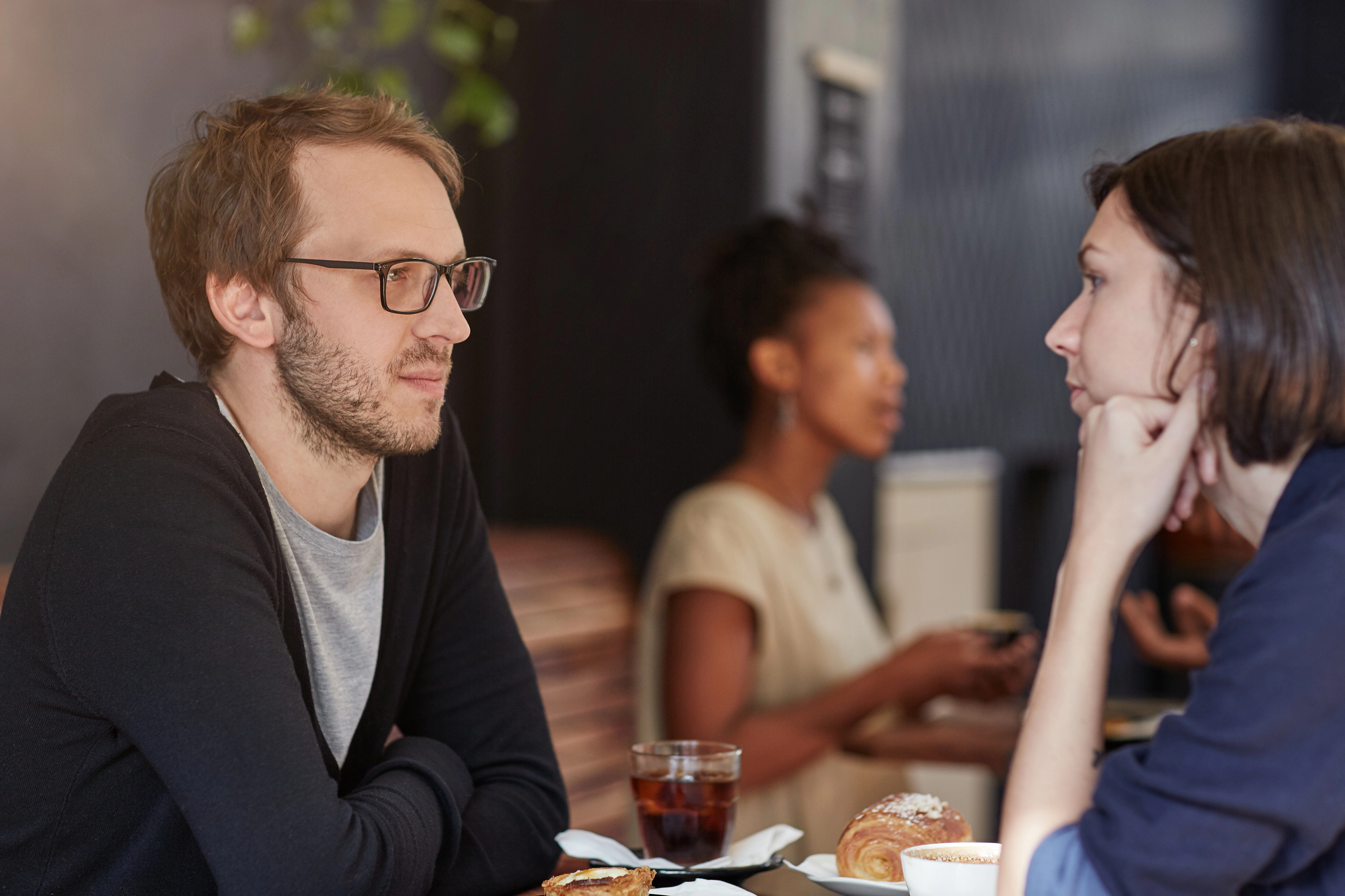 A man with glasses and a beard, dressed in a black cardigan and gray shirt, sits at a cafe table conversing with a woman who has short dark hair and is resting her chin on her hand. Two other people are blurred in the background, engaged in their own conversation.