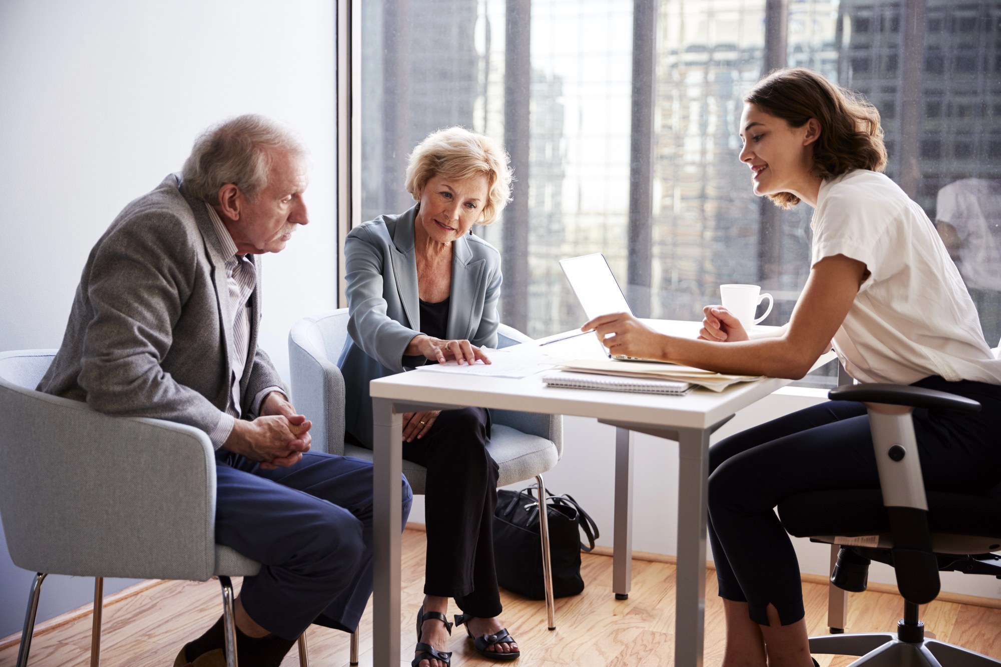 Three people sit around a table in an office. An older man and woman are on one side, looking at a young woman on the other side, who is holding a laptop. Papers and a coffee cup are on the table, and large windows are in the background.