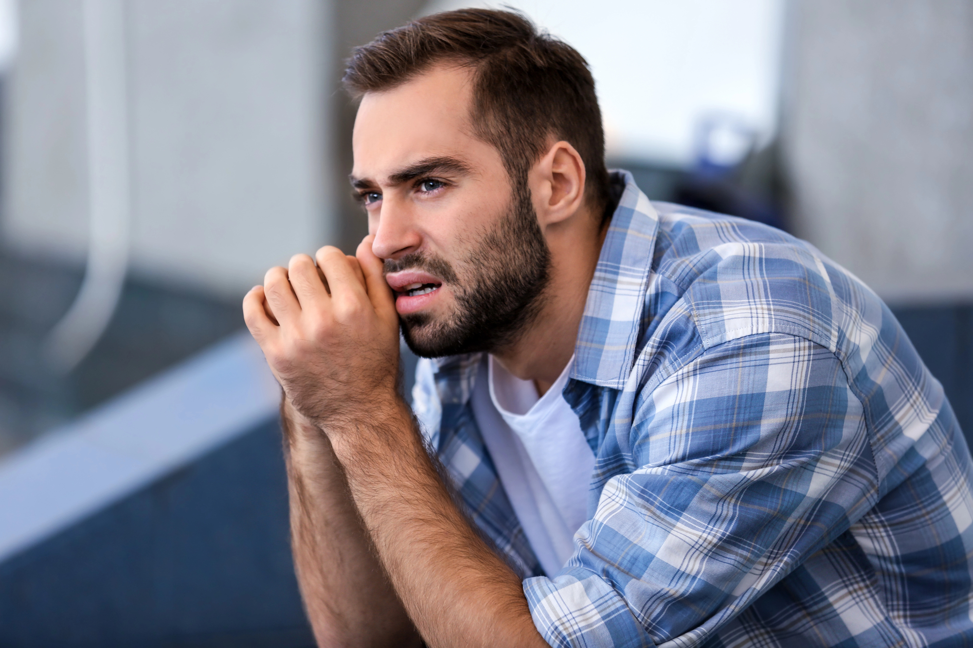 A man with a beard and short hair sits pensively, resting his chin on his hands. He is wearing a blue and white plaid shirt over a white t-shirt. The background is blurred, suggesting an indoor setting.