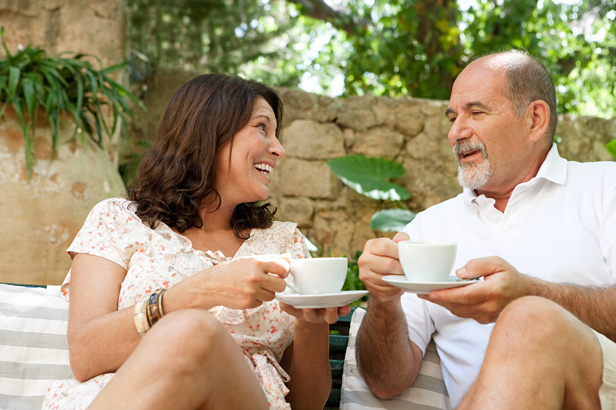 A woman and a man are sitting outdoors, smiling at each other while holding white teacups. They are surrounded by greenery and a stone wall. The woman wears a floral dress, and the man is dressed in a white polo shirt.