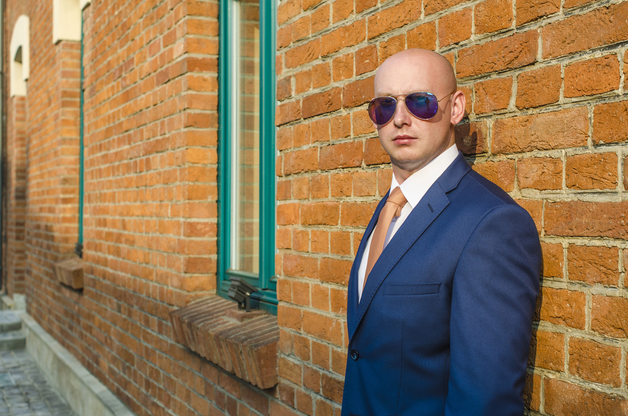 A man in a blue suit and orange tie leans against a brick wall. He is wearing sunglasses and has a serious expression. The wall features green-trimmed windows and the scene is set outdoors.