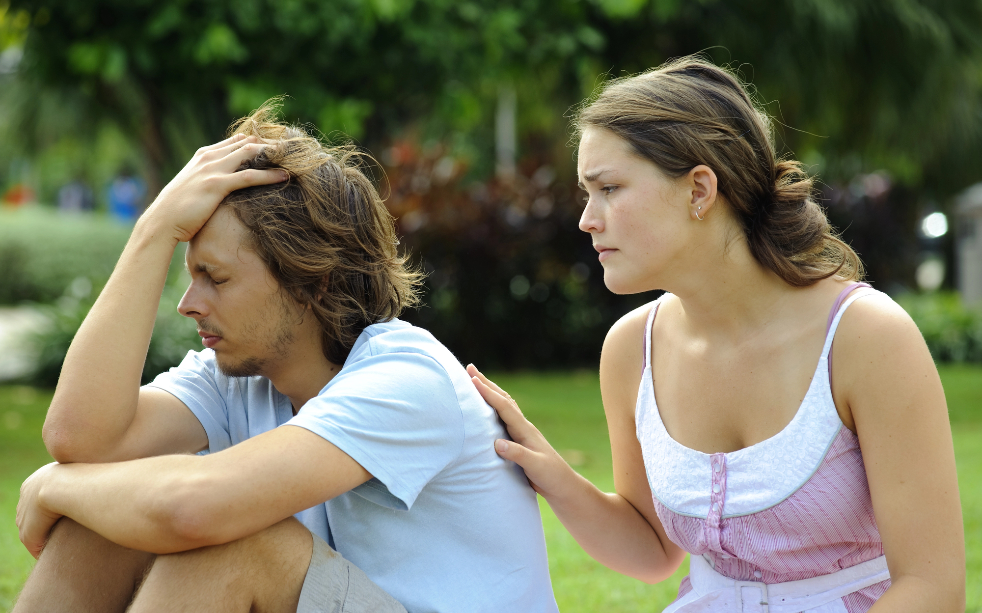 A woman sits beside a man in a park. The man, appearing upset, rests his head in his hand. The woman, wearing a pink and white dress, gently touches his back, offering comfort. Greenery and trees are visible in the background.