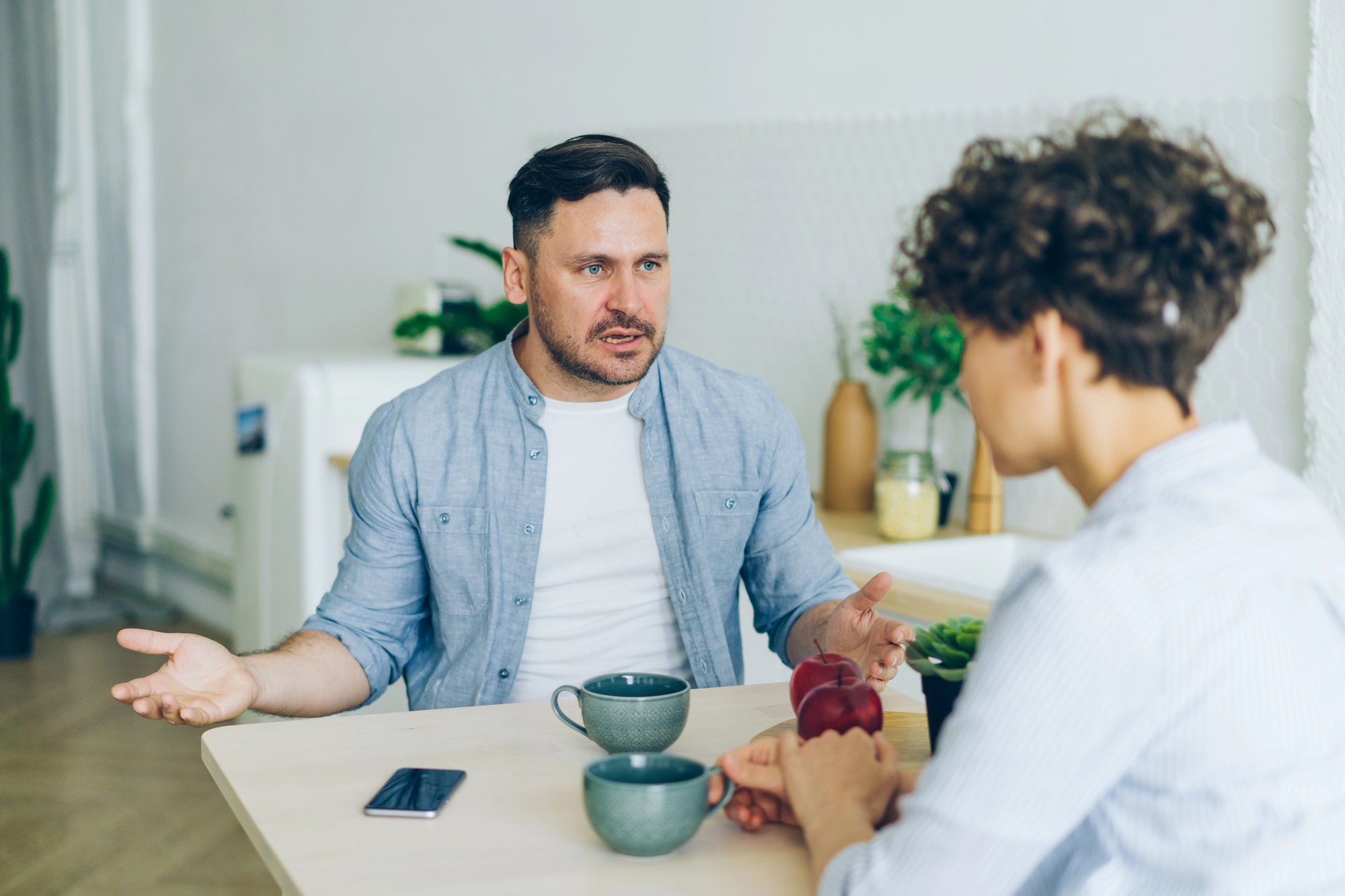 Two people sit at a table having a conversation. One gestures with their hands, while the other listens. They have mugs in front of them and a smartphone is placed on the table. The background includes potted plants and a small kitchen area.