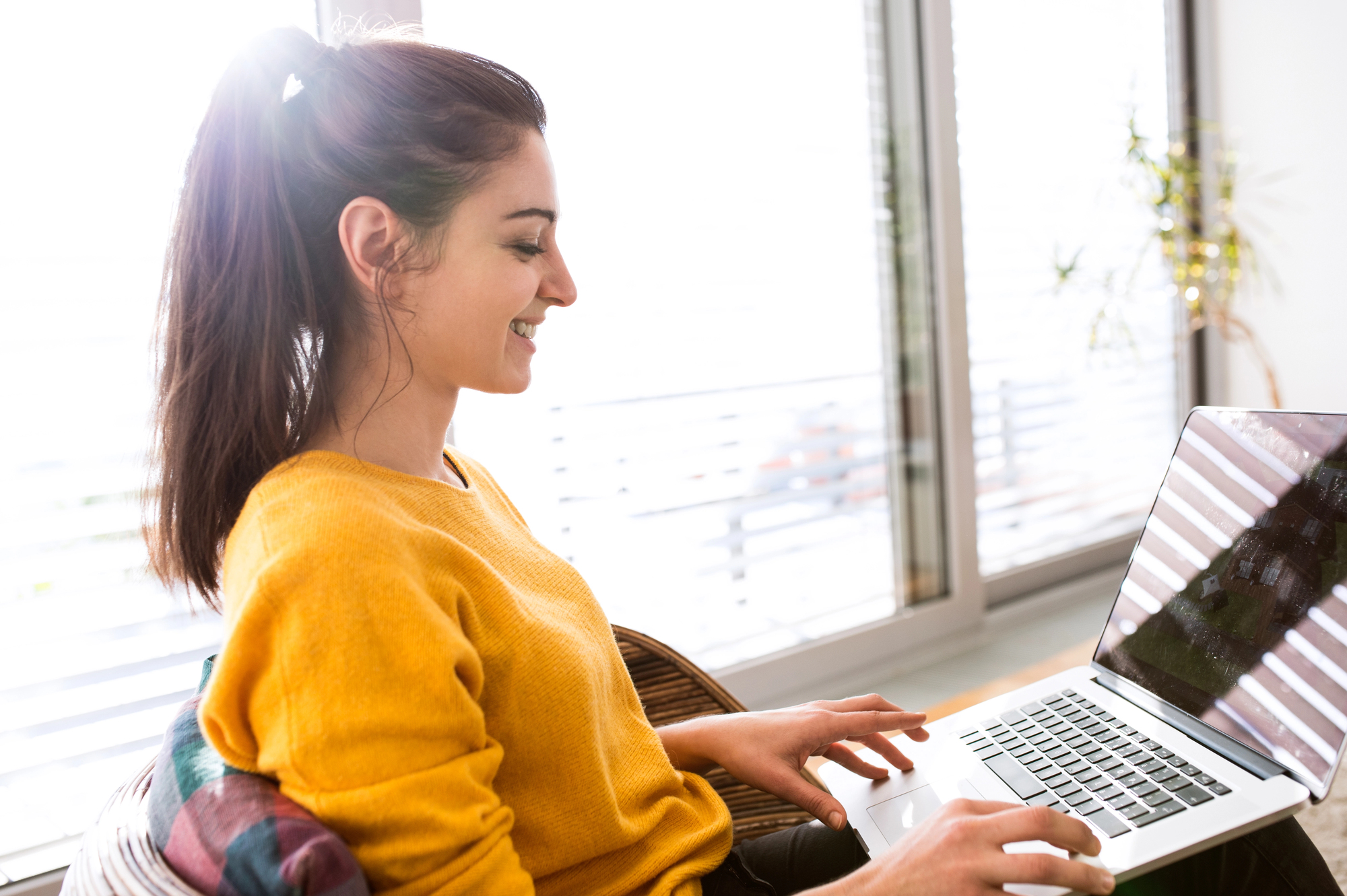 A woman in a yellow sweater is sitting and smiling while using a laptop. She is positioned near large windows with blinds, allowing natural light to illuminate the space. A small plant is visible in the background.