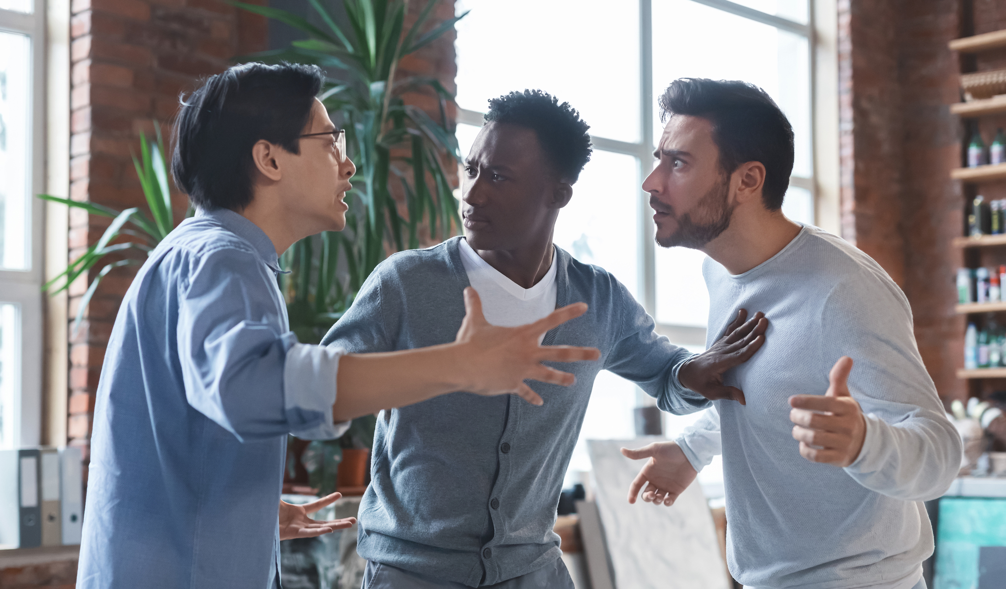 Three men appear to be in a heated discussion in what looks like a modern office setting. One man on the left gestures with open hands, the man in the middle looks concerned, and the man on the right points his finger, seeming defensive. Sunlight streams through large windows.