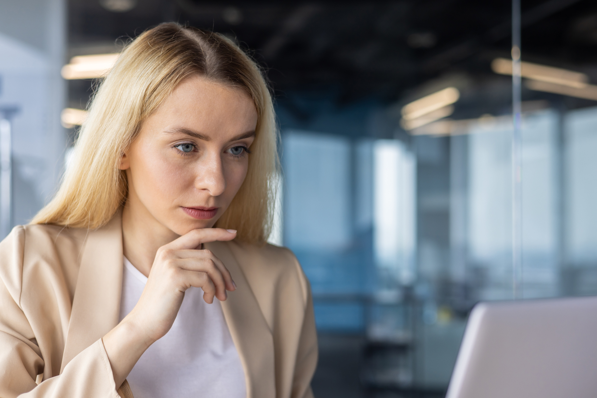 A woman with long blonde hair is sitting in a modern office, looking intently at a laptop screen. She is wearing a beige blazer over a white top. Her hand is near her chin, and there are large windows in the background.