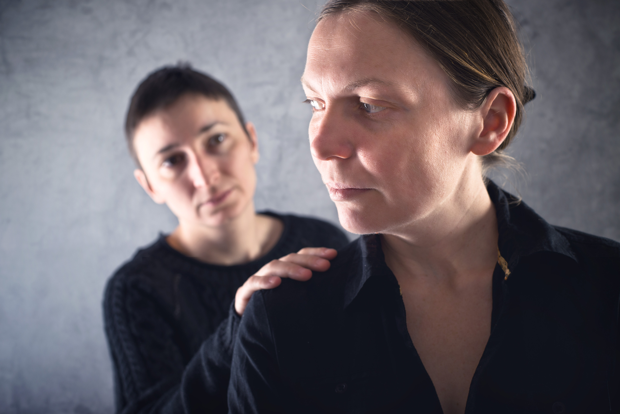 Two people are in the image. The person in the foreground, looking away, wears a black shirt. The person in the background has a short hairstyle and a hand on the other's shoulder, appearing empathetic. The background is a neutral gray.