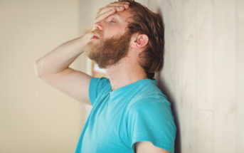 A man with a beard stands against a wooden wall, wearing a blue shirt. He has his eyes closed and his hand on his forehead, appearing stressed or frustrated.