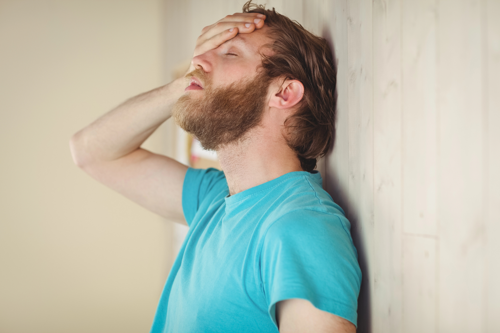 A man with a beard stands against a wooden wall, wearing a blue shirt. He has his eyes closed and his hand on his forehead, appearing stressed or frustrated.