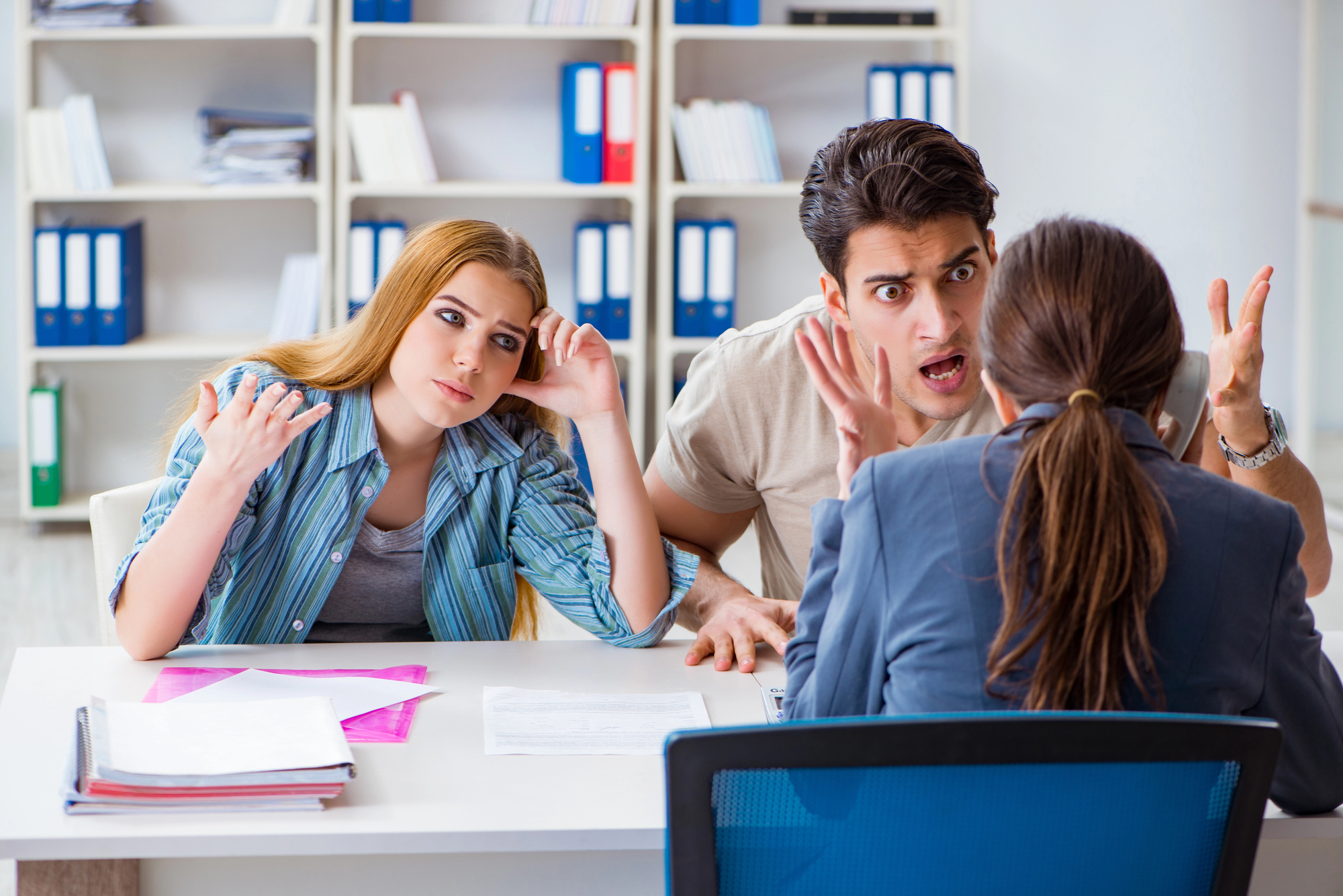 A frustrated couple is sitting across from a professional-looking woman in an office setting. The man is gesturing with his hands, appearing upset, while the woman beside him rests her head on her hand, looking tired. Shelves with binders are in the background.