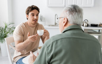 A young man holding a coffee cup talks to an older man seated at a table in a bright kitchen. The young man has an animated expression, while the older man listens intently. There are kitchen utensils and plants in the background.