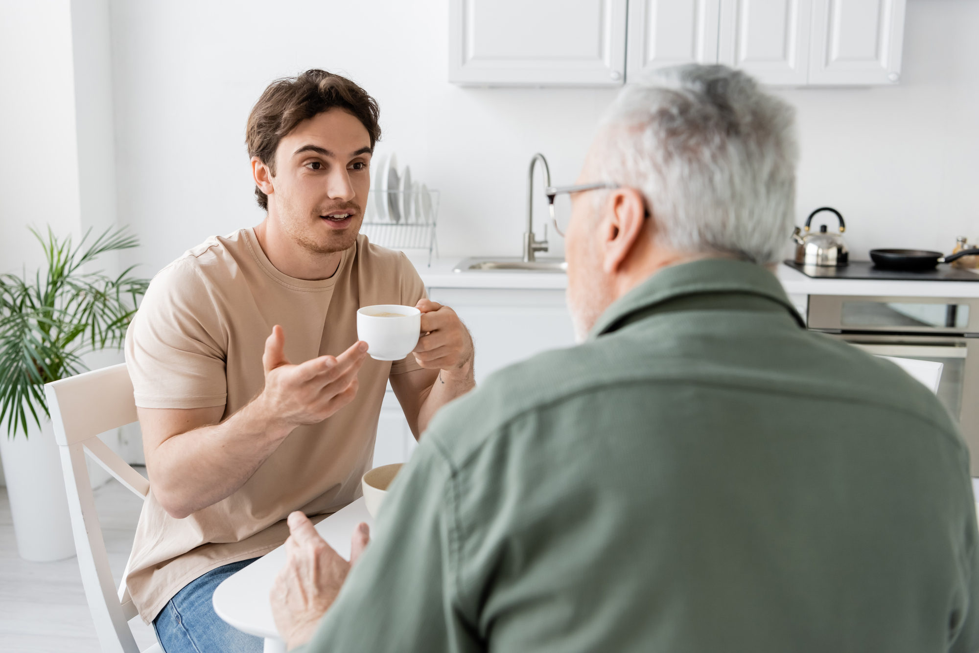 A young man holding a coffee cup talks to an older man seated at a table in a bright kitchen. The young man has an animated expression, while the older man listens intently. There are kitchen utensils and plants in the background.