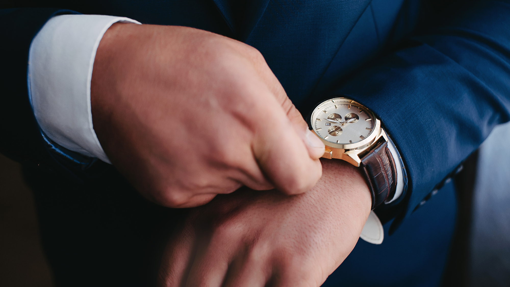 Close-up of a person in a blue suit adjusting a gold wristwatch with a brown leather strap on their left wrist.