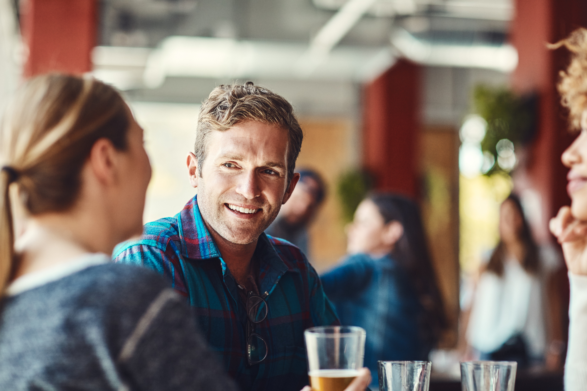 A man in a plaid shirt holds a drink and smiles while sitting at a table conversing with two people in a casual setting. Blurred figures and soft lighting indicate a relaxed and social atmosphere.