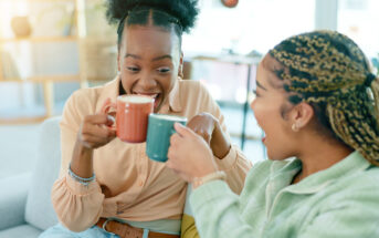 Two people sit on a couch, smiling and laughing while clinking their mugs together. One person wears an orange shirt and holds a red mug, and the other wears a green shirt and holds a blue mug. The background shows a cozy and well-lit room.