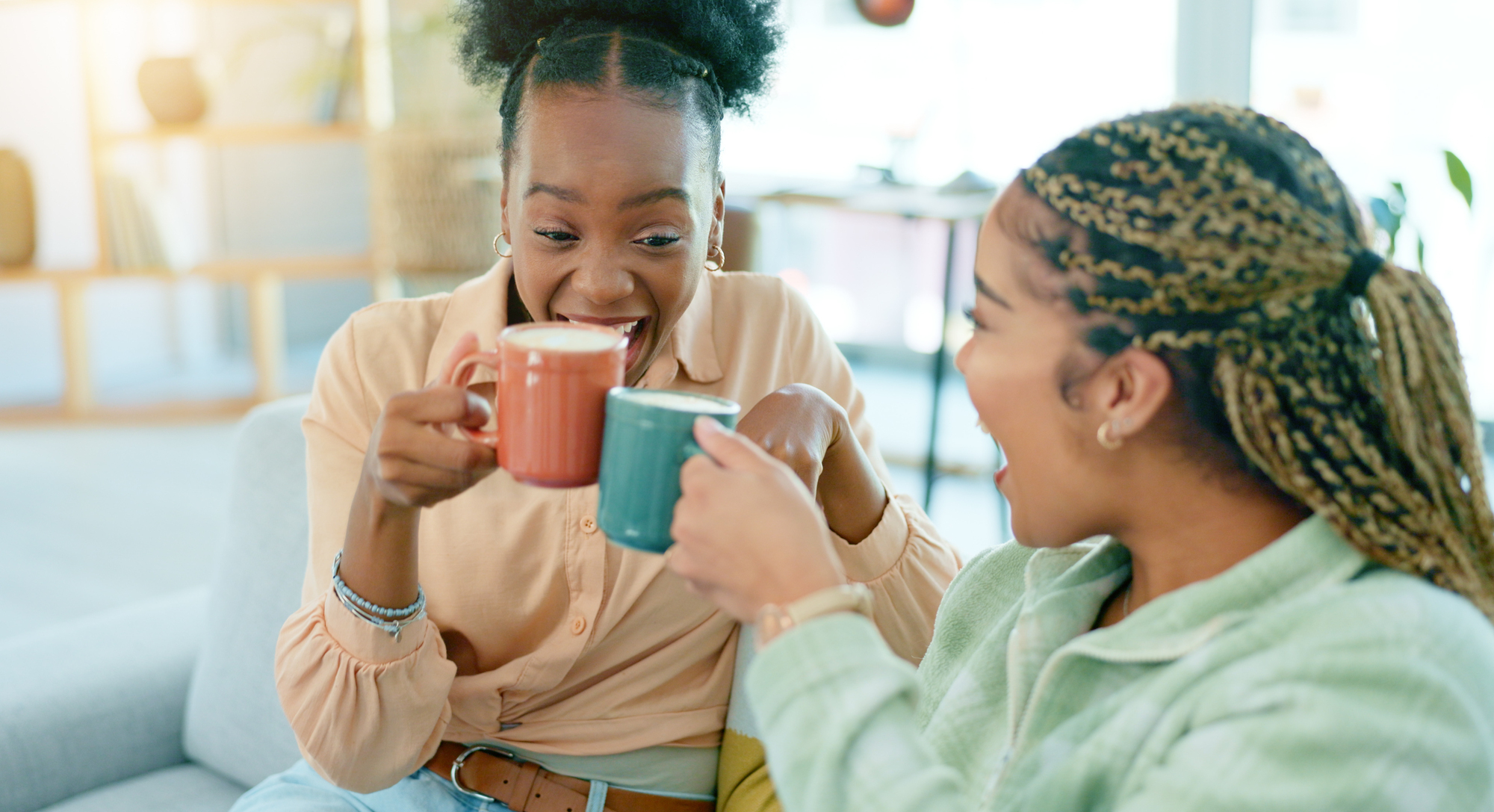 Two people sit on a couch, smiling and laughing while clinking their mugs together. One person wears an orange shirt and holds a red mug, and the other wears a green shirt and holds a blue mug. The background shows a cozy and well-lit room.