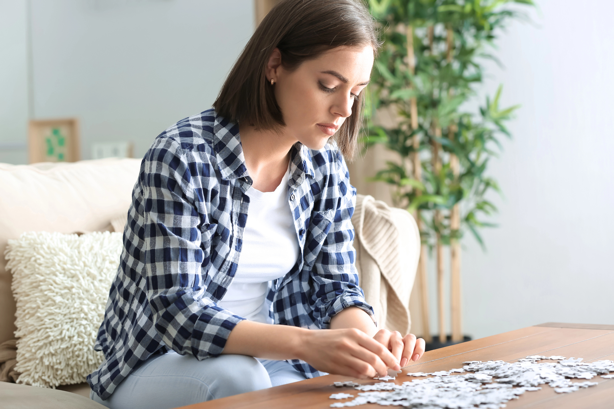 A woman with shoulder-length brown hair, wearing a blue and white checkered shirt, is sitting on a beige sofa and assembling a puzzle on a wooden table. A leafy green plant is in the background.