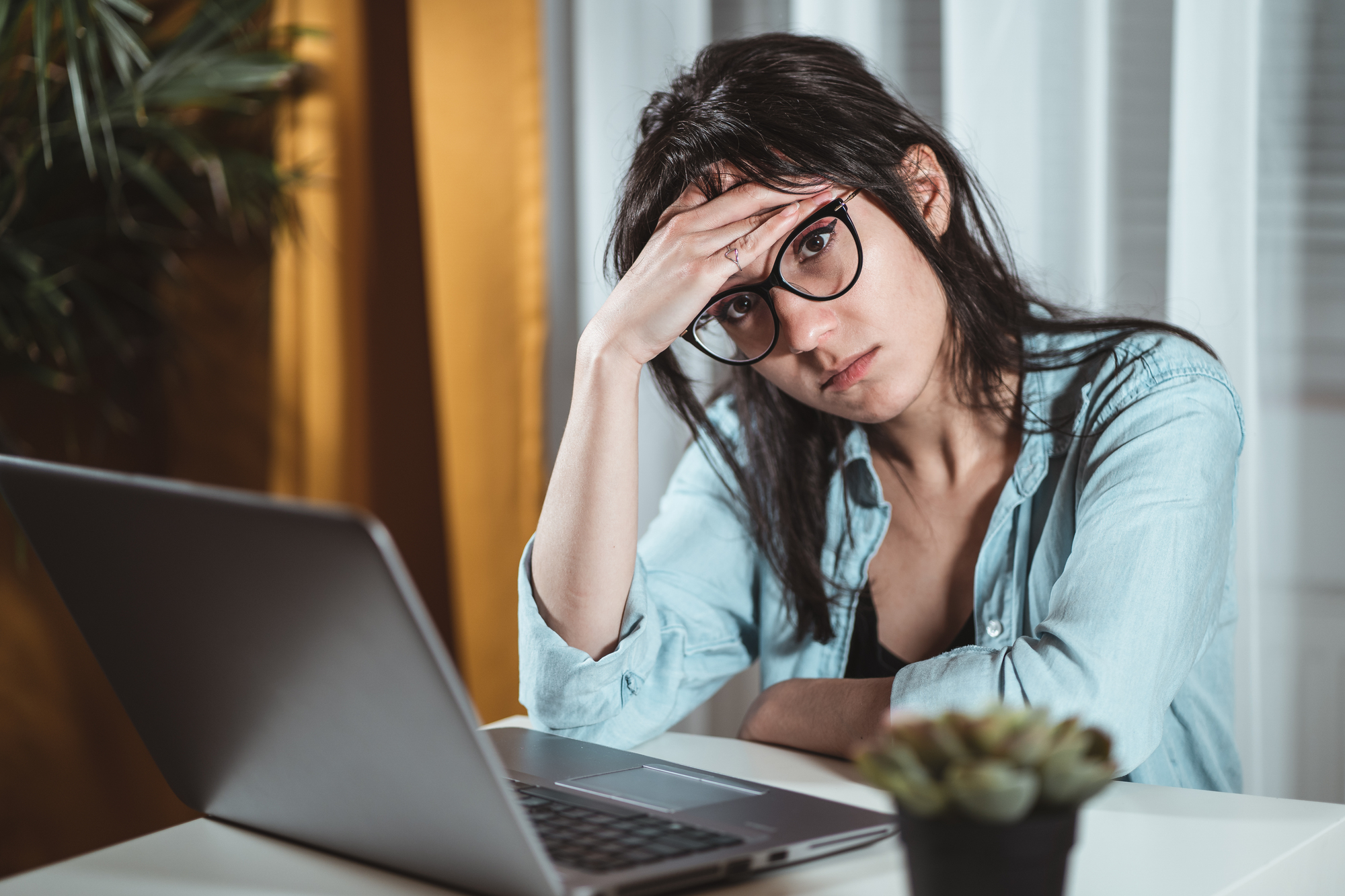 A woman with glasses and dark hair sits at a desk with her open laptop in front of her. She rests her head on her hand, appearing stressed or tired. A small potted plant is also on the desk. Light filters through window blinds in the background.