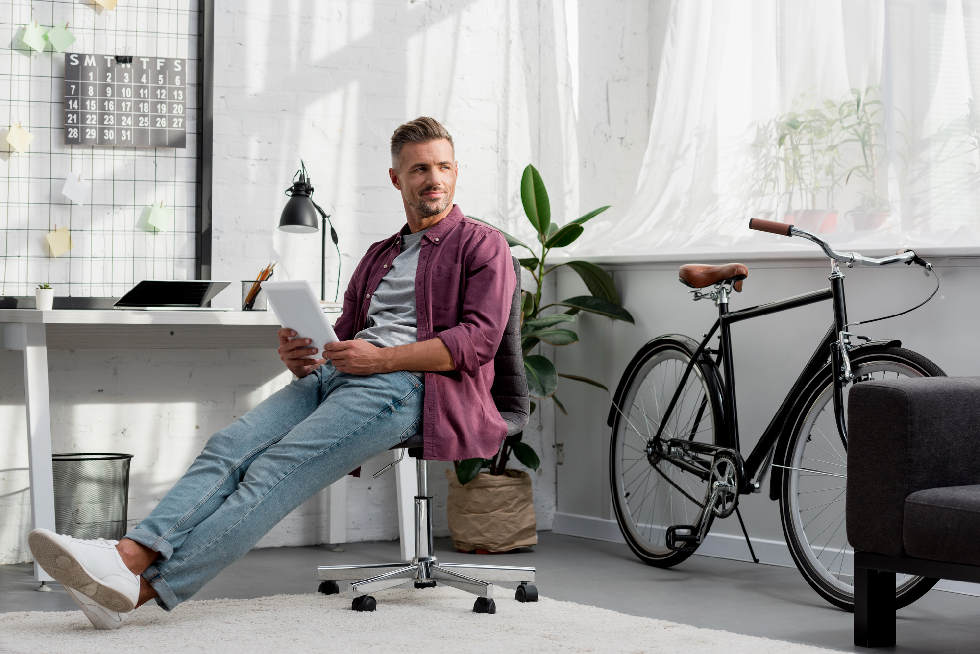 A man sits casually on a swivel chair, holding a tablet, in a stylish office with a bicycle beside him. The room features a modern desk with a laptop, a lamp, a wall calendar, and a large window with sheer curtains.