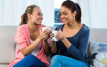 Two women sit on a couch, smiling and holding mugs. One is wearing a pink cardigan over a white top, and the other is in a blue sweater. They appear to be enjoying a pleasant conversation in a bright room with white curtains and a cushion beside them.