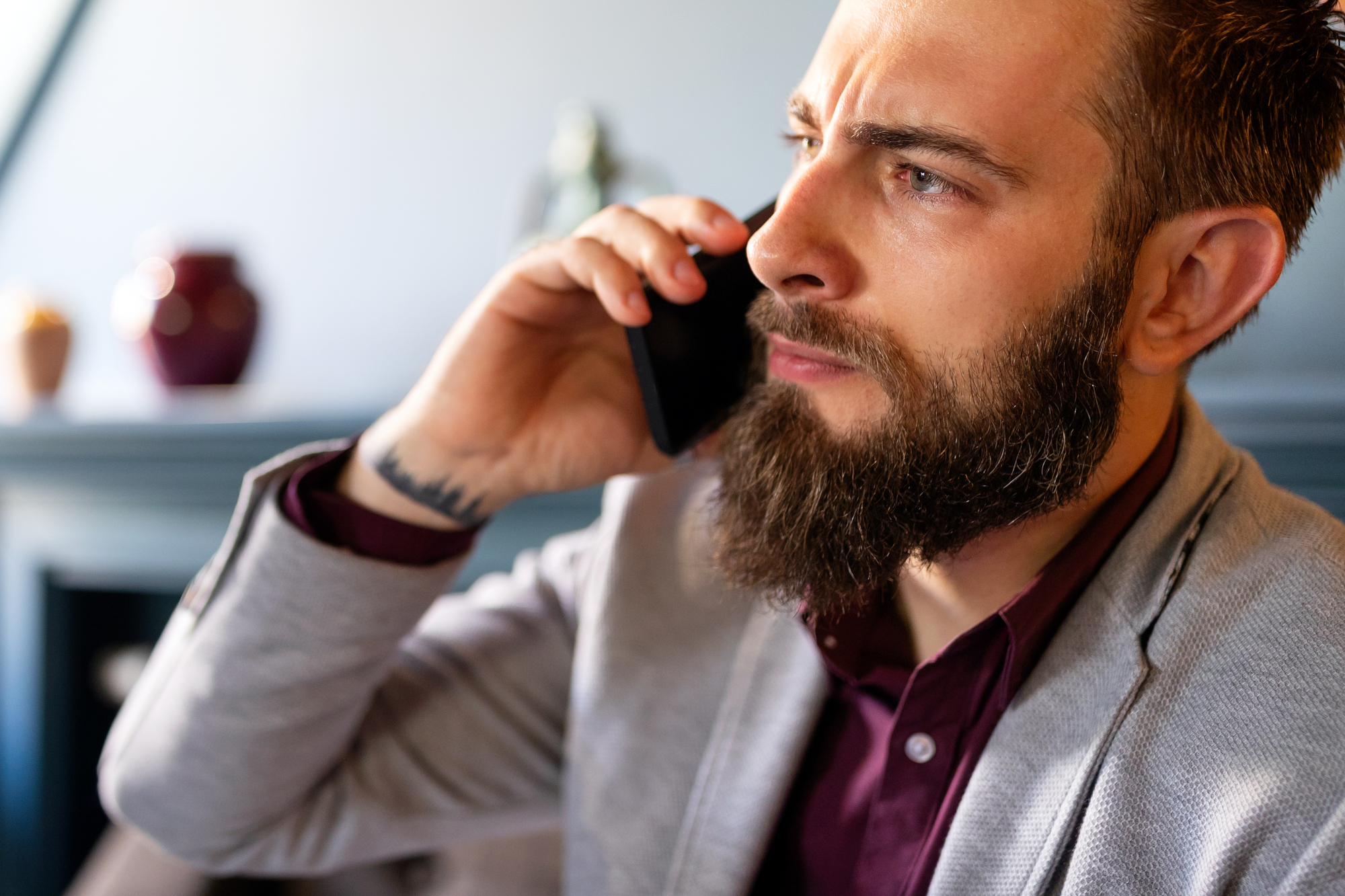 A man with a beard and wearing a gray blazer and maroon shirt holds a smartphone to his ear. He appears to be engaged in a serious phone conversation. Blurred background items include a pot and abstract shapes.