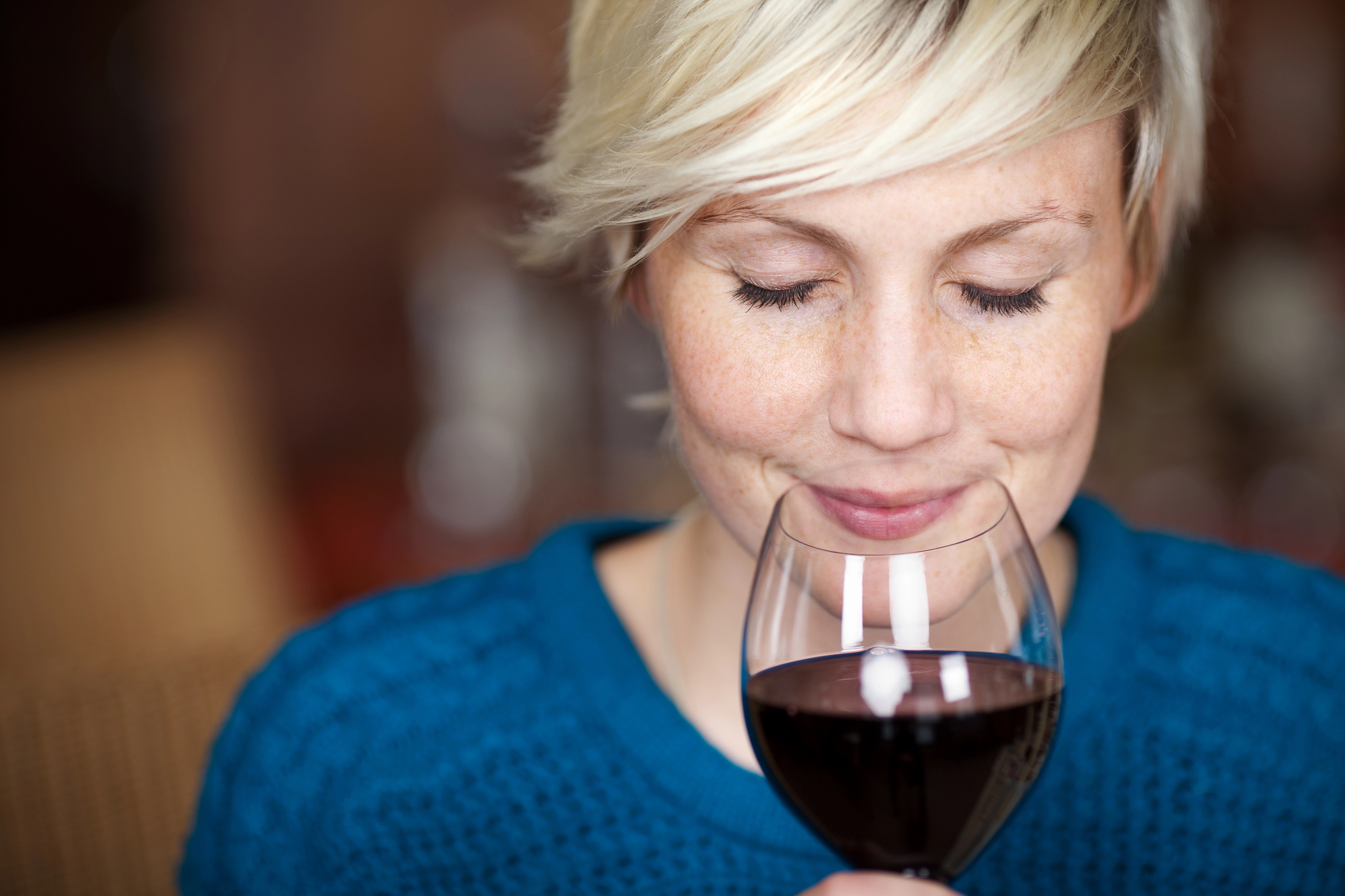 A person with short blonde hair is holding a glass of red wine near their face. Their eyes are closed and they are smiling slightly, as if enjoying the aroma. They are wearing a blue sweater. The background is softly blurred.