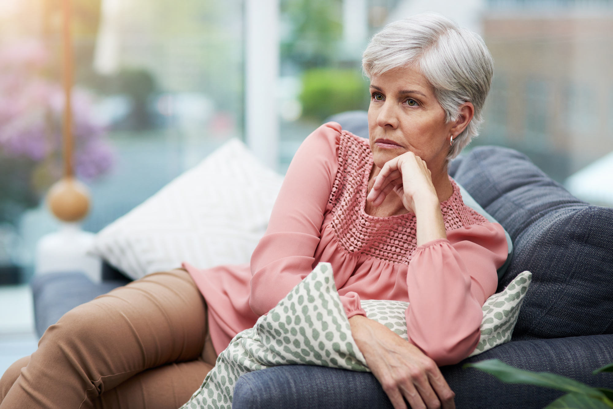 An older woman with short gray hair, wearing a pink blouse and beige pants, sits on a couch looking contemplatively out a window. She rests her chin on her hand, with decorative pillows behind her in a softly lit room.