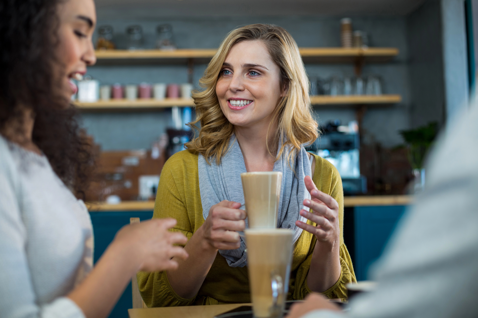 Three people sit at a café table enjoying drinks. A woman in the center, with blond hair and a grey scarf, smiles while holding a glass of latte. Another person on the left, partially visible, talks animatedly. Shelves with jars and cups are in the background.