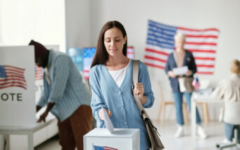 A woman in a blue shirt is casting her vote at a polling station. Behind her, a person is voting, and two others are standing next to a table with papers. An American flag is displayed on the wall.