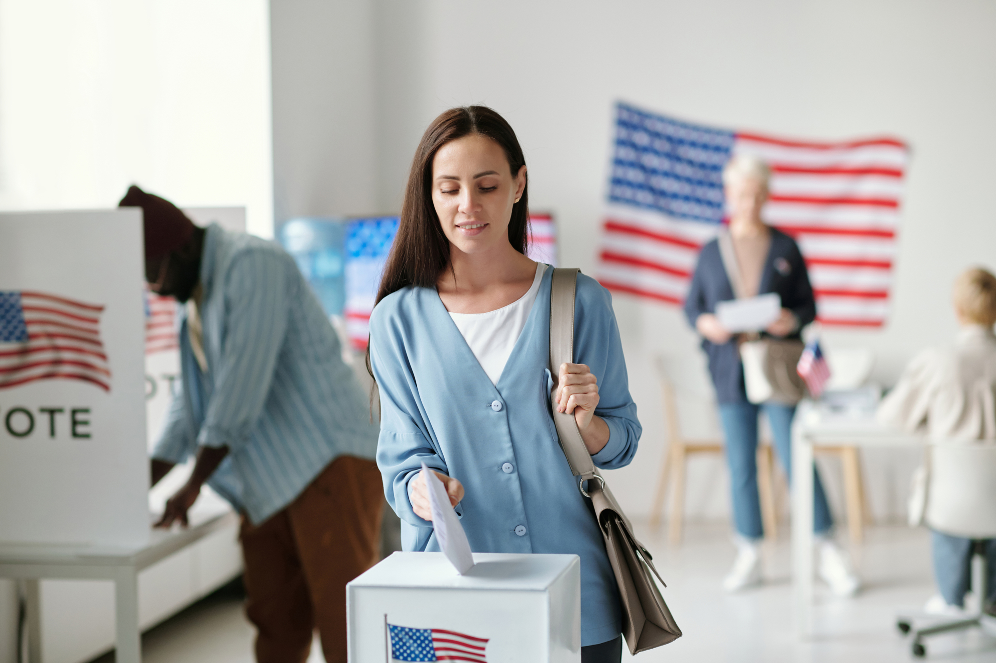 A woman in a blue shirt is casting her vote at a polling station. Behind her, a person is voting, and two others are standing next to a table with papers. An American flag is displayed on the wall.