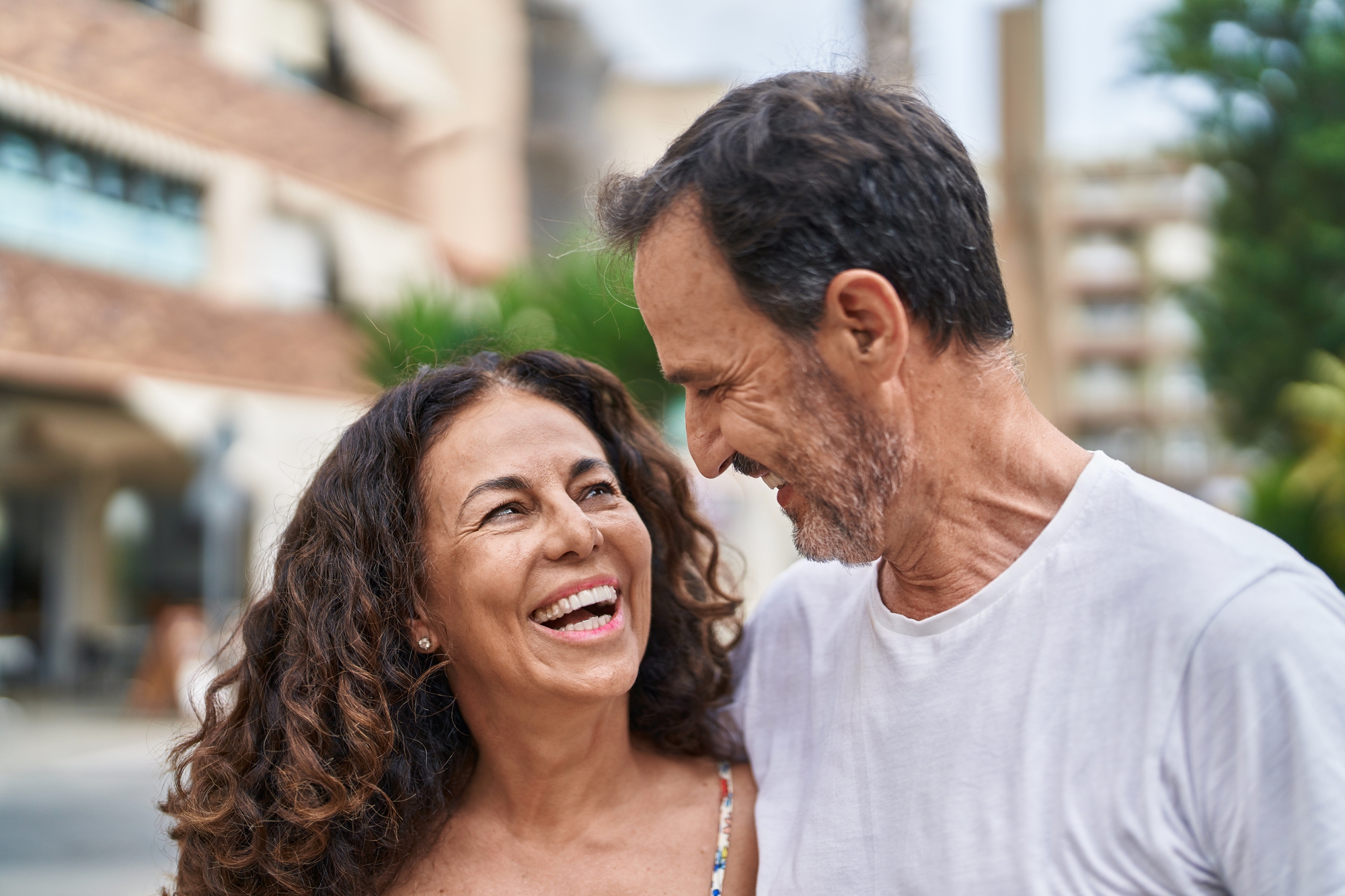 A joyful couple embraces outdoors, smiling at each other. The woman has long curly hair, and the man has short hair and a beard. The background features a blurred view of a building and greenery.