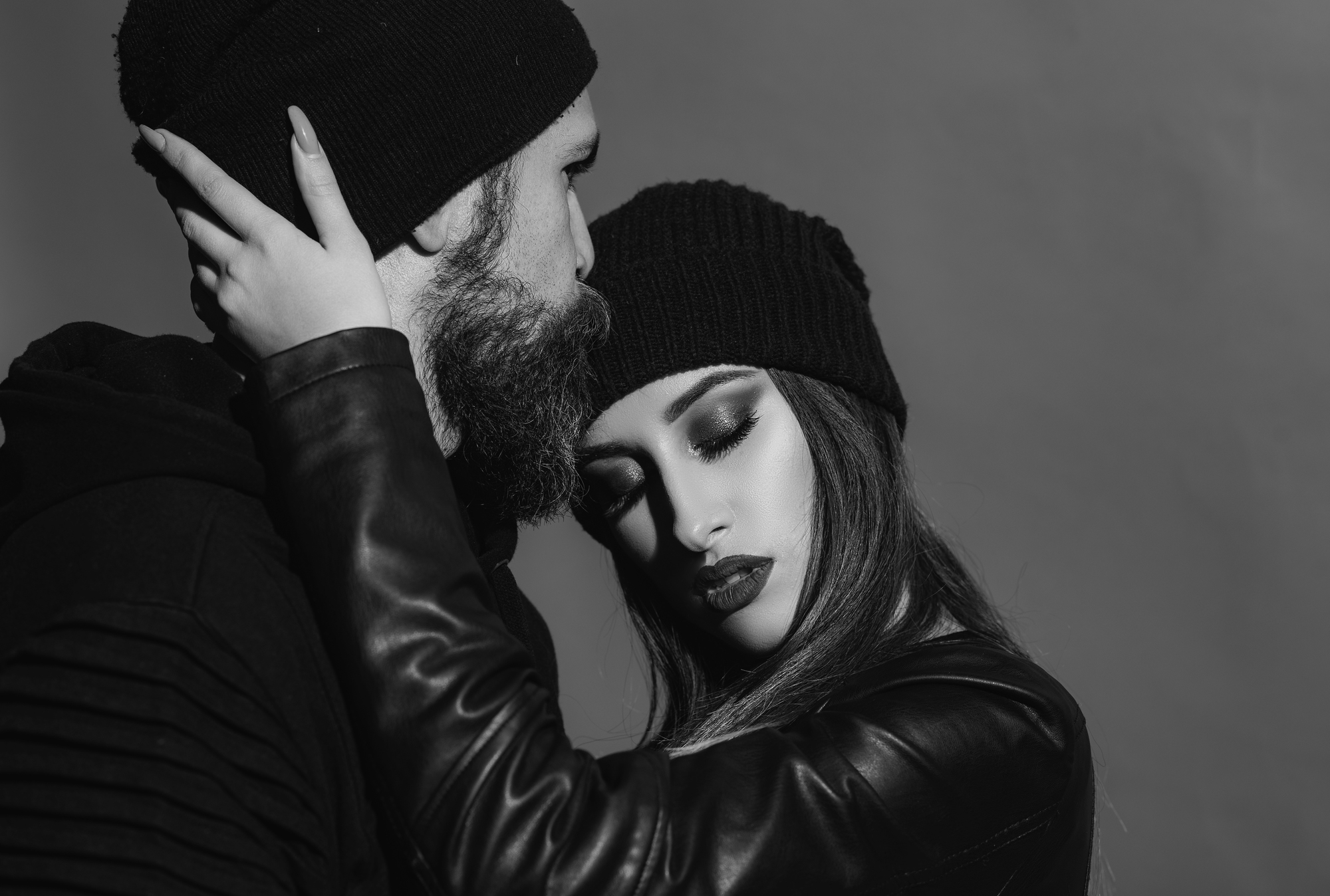 Black and white photo of a man kissing a woman's forehead. Both wear dark beanies and leather jackets. The woman has long hair and closed eyes, showing an expression of calm. The background is a simple gradient.