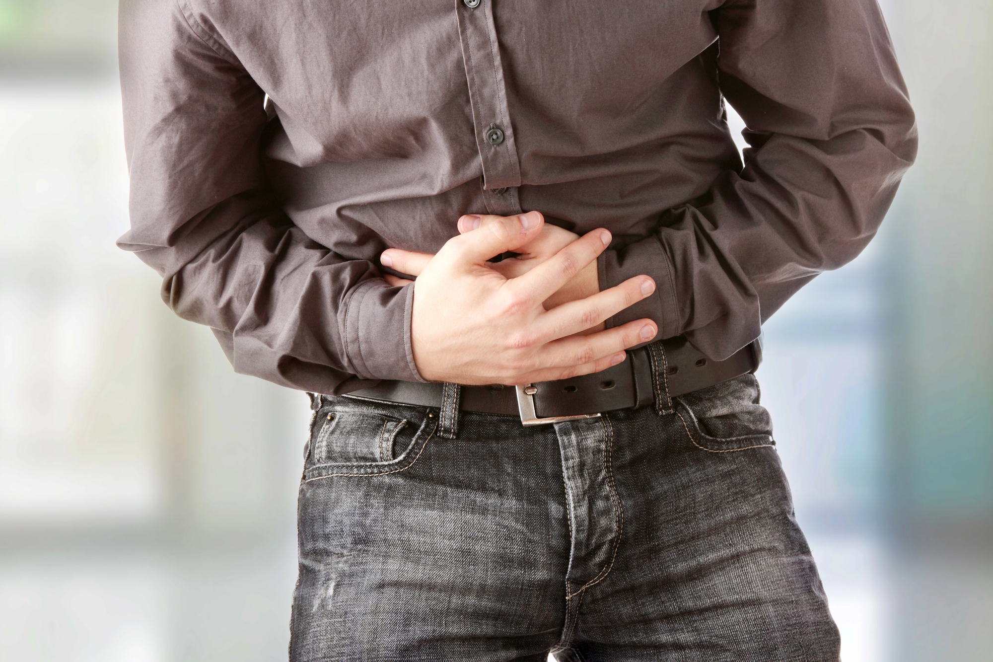 A person is clutching their stomach with both hands, appearing to experience abdominal pain. They are wearing a gray button-up shirt and blue jeans with a black belt. The background is blurred, putting focus on the individual's discomfort.