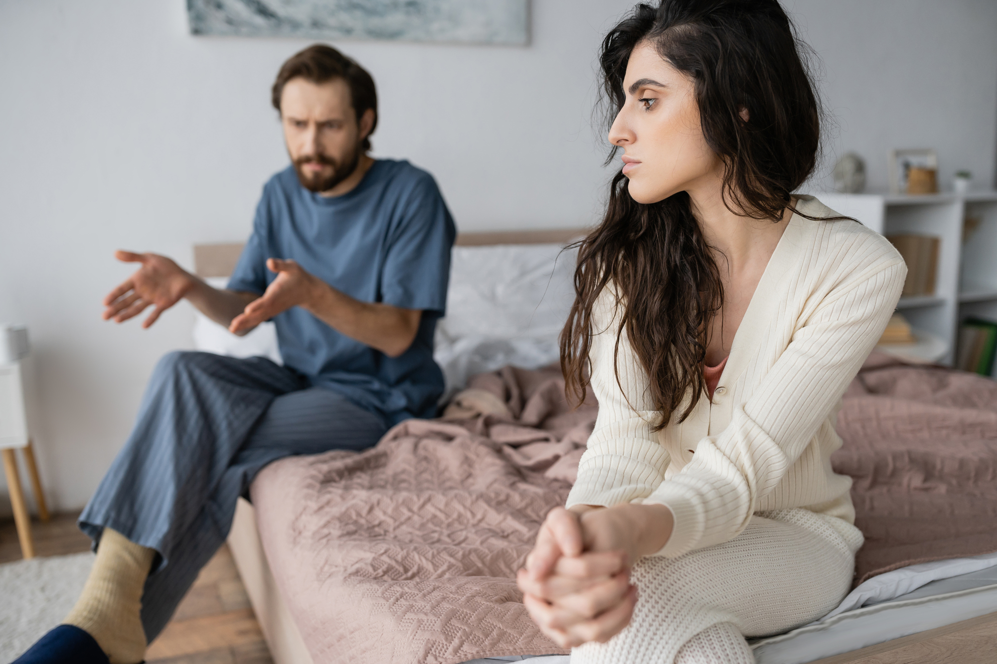 A man and a woman are sitting on a bed in a bedroom. The man, dressed in a blue shirt and gray pants, appears to be gesturing and speaking with a serious expression. The woman, wearing a light-colored cardigan and pants, looks away, appearing upset or thoughtful.