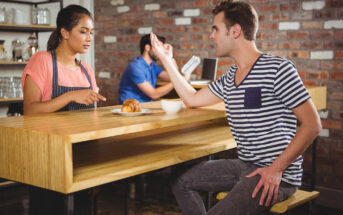 A man sitting at a wooden counter in a café is gesturing and speaking to a waitress in an apron. A pastry and coffee are in front of him. Another person is seated in the background, reading a menu. A brick wall is visible behind them.