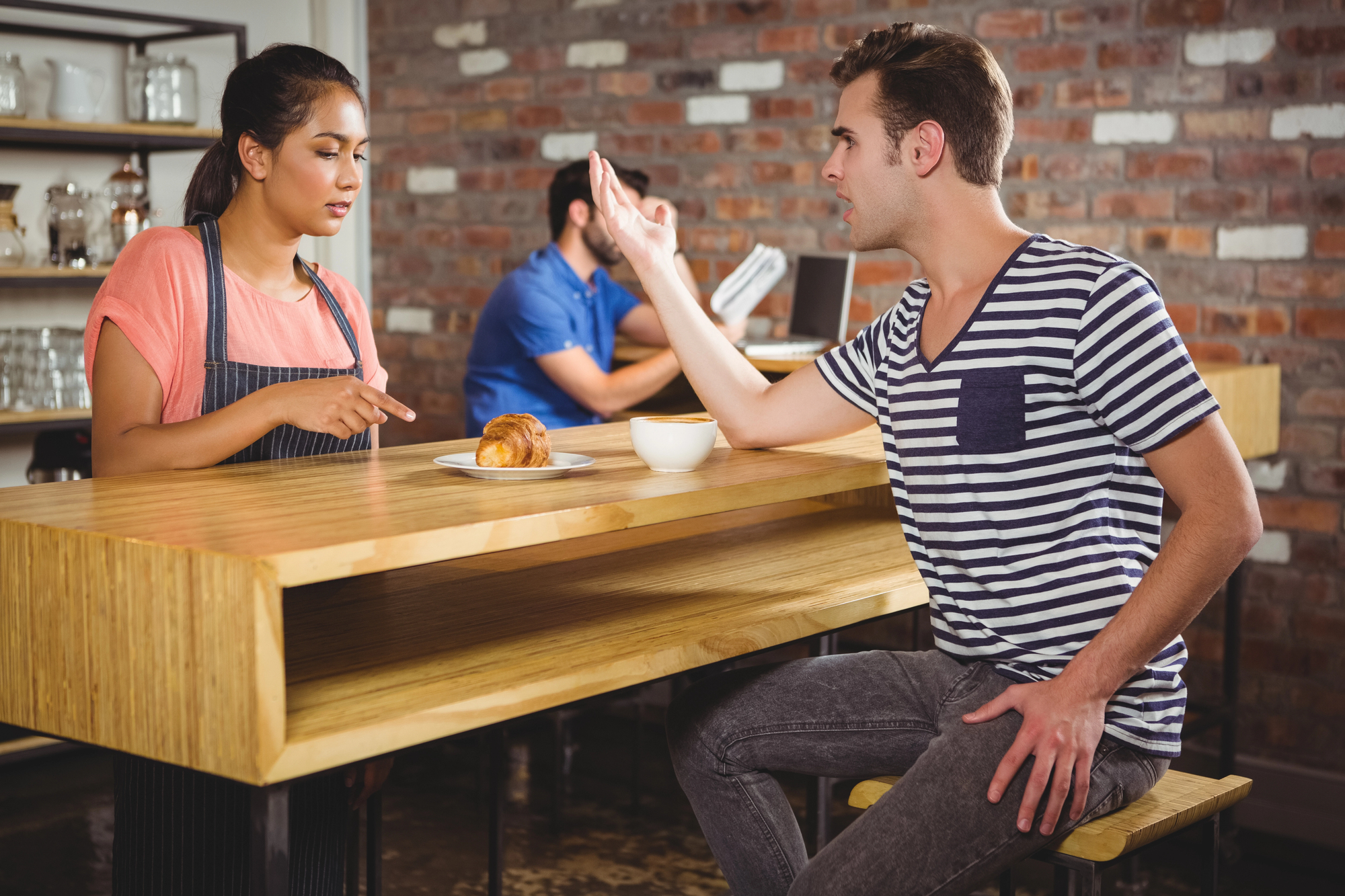 A man sitting at a wooden counter in a café is gesturing and speaking to a waitress in an apron. A pastry and coffee are in front of him. Another person is seated in the background, reading a menu. A brick wall is visible behind them.