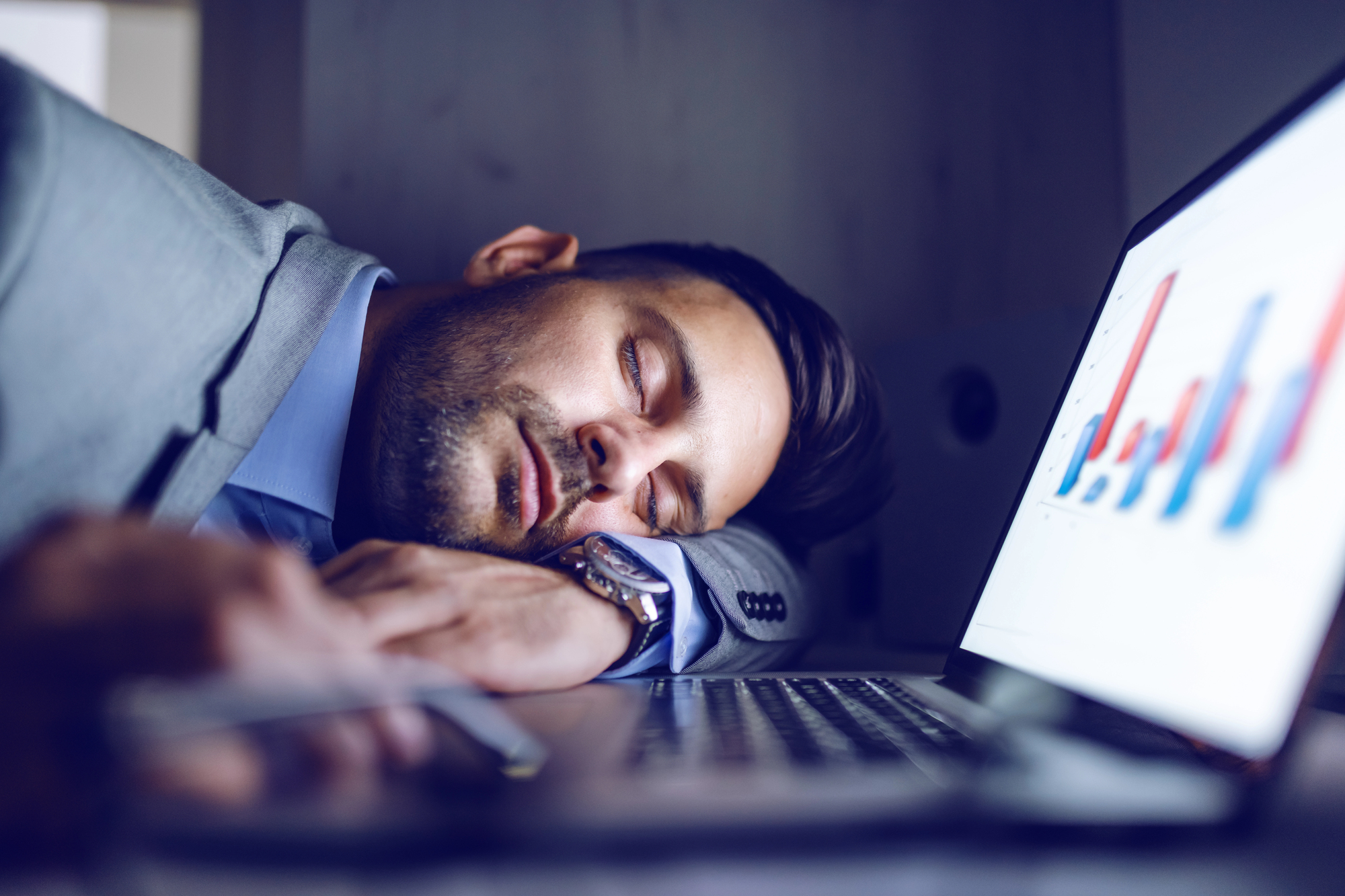 A man in a suit is asleep at a desk with his head resting on his arms. In front of him, a laptop displays a graph with blue and red bars. A pen and smartphone are also on the desk.