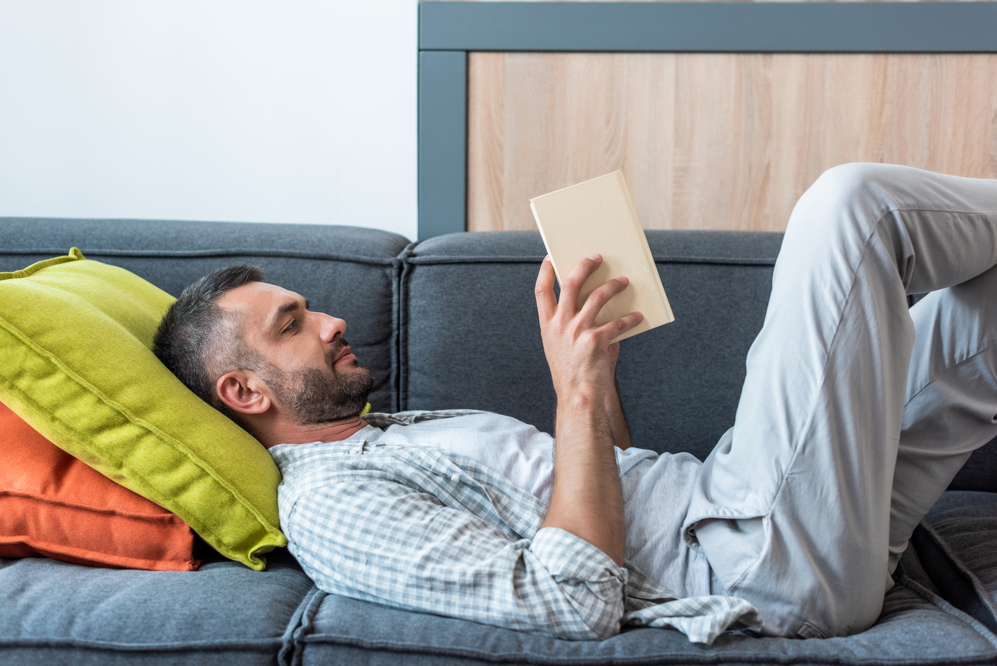 A man with a beard is lying on a gray sofa, propped up against green and orange cushions, reading a book. He is wearing a casual, light-colored outfit and appears relaxed in a comfortable, well-lit room.