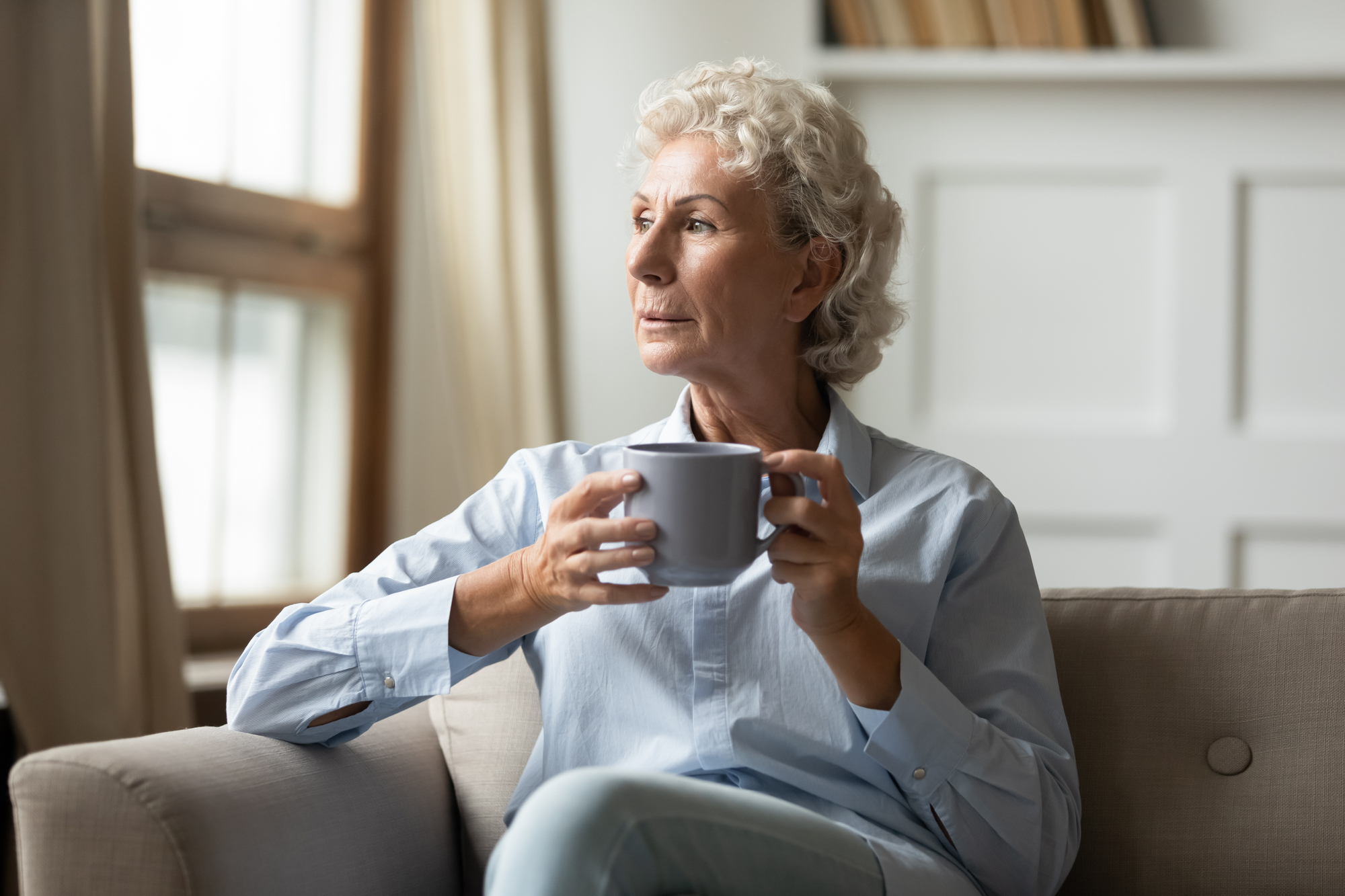 An elderly woman with short, curly gray hair sits on a sofa holding a large mug, gazing thoughtfully out a window. She is wearing a light blue shirt, and the room is softly lit with a bookshelf in the background.