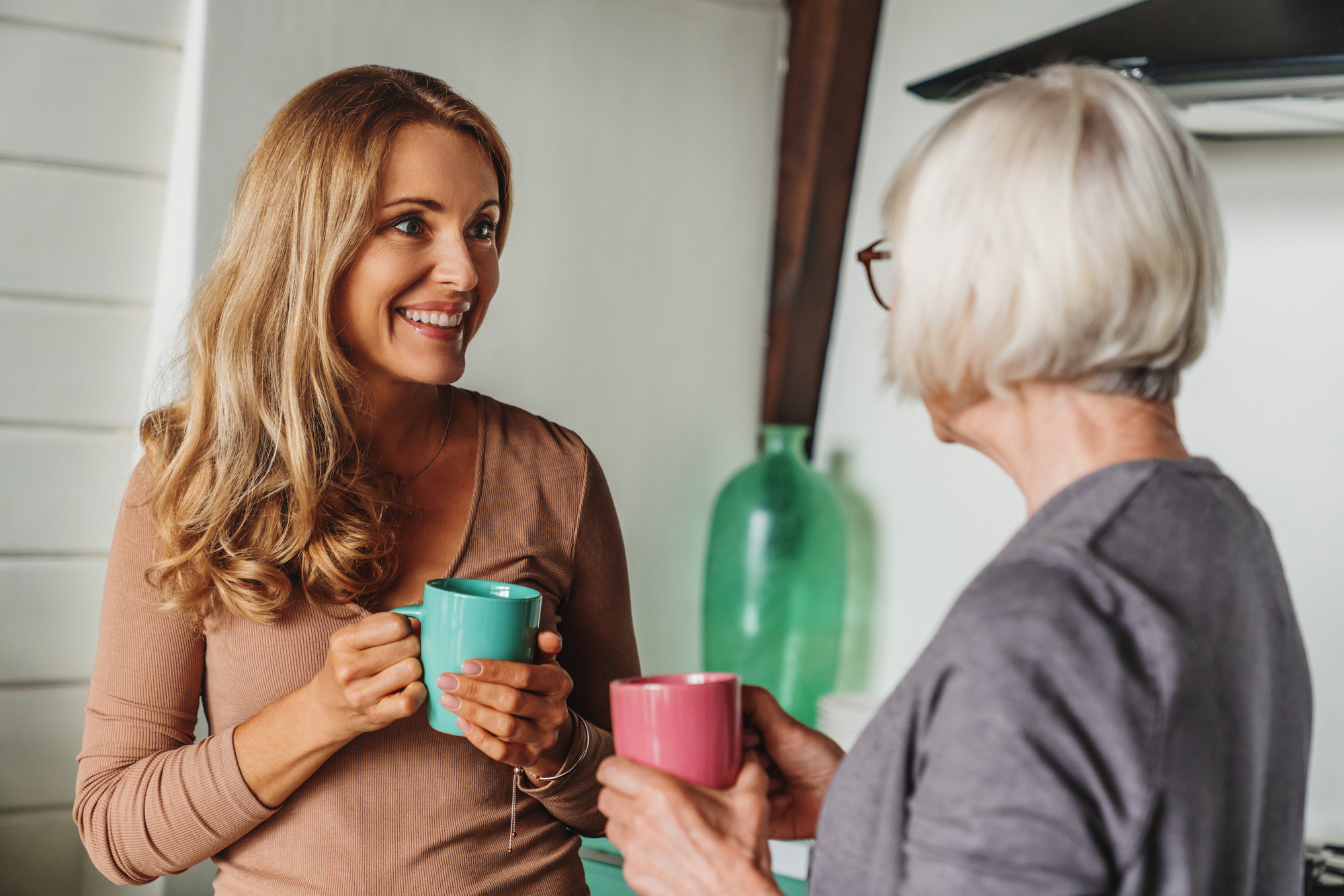 A young woman with long hair smiles while talking to an older woman with short gray hair. They are holding mugs in a kitchen setting, with a large green bottle in the background.